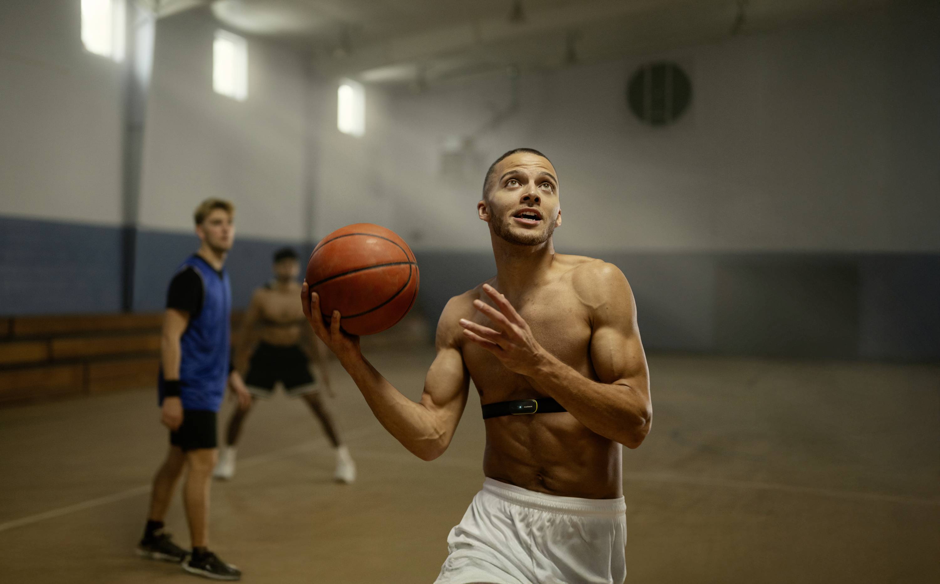 Un homme en tenue de sport saute avec un ballon de basket dans un gymnase. Deux autres personnes sont visibles en arrière-plan.