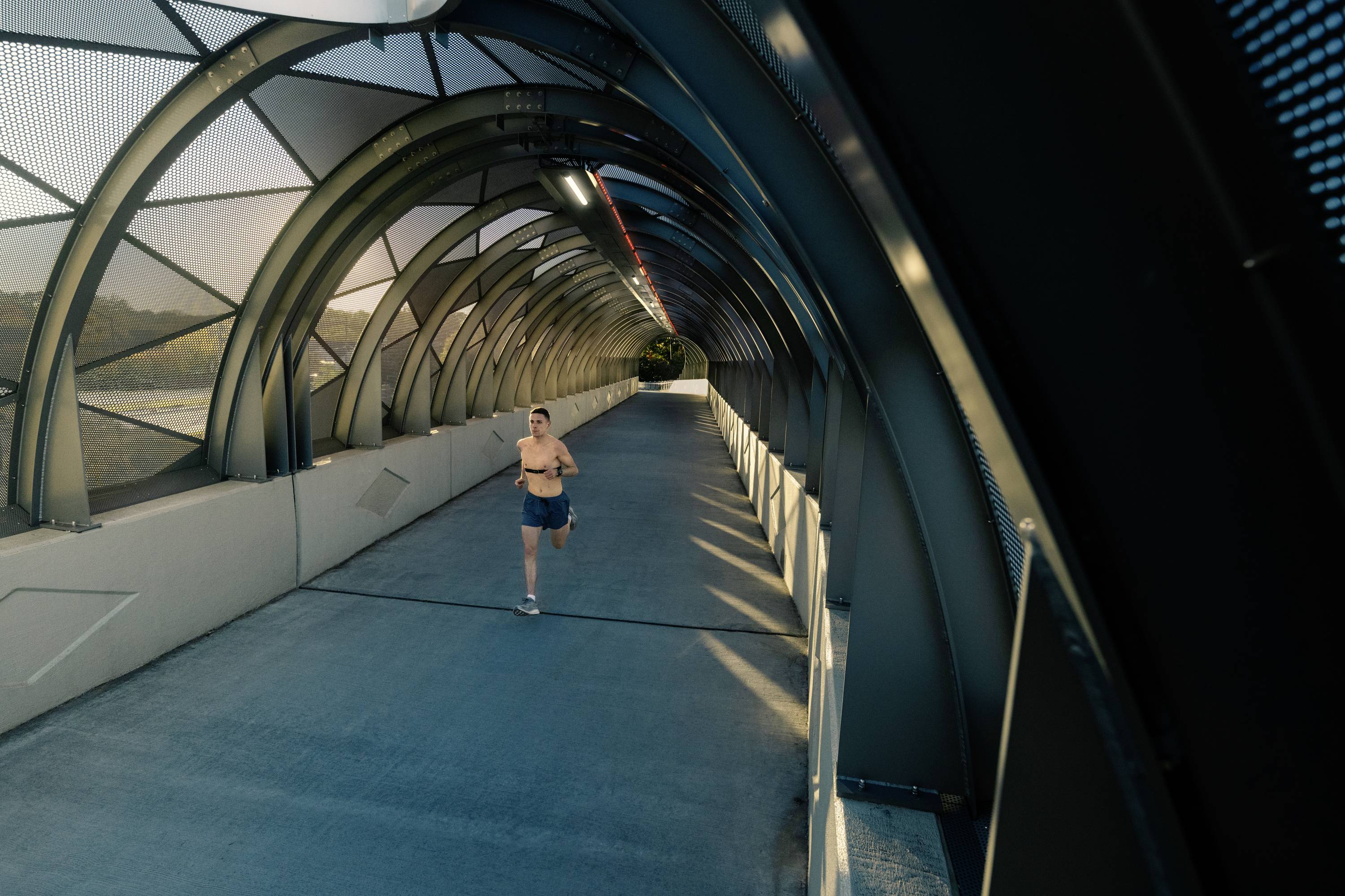 Une personne fait du jogging dans un tunnel au design futuriste avec une architecture en forme d'arche. La lumière pénètre à travers les structures ouvertes.