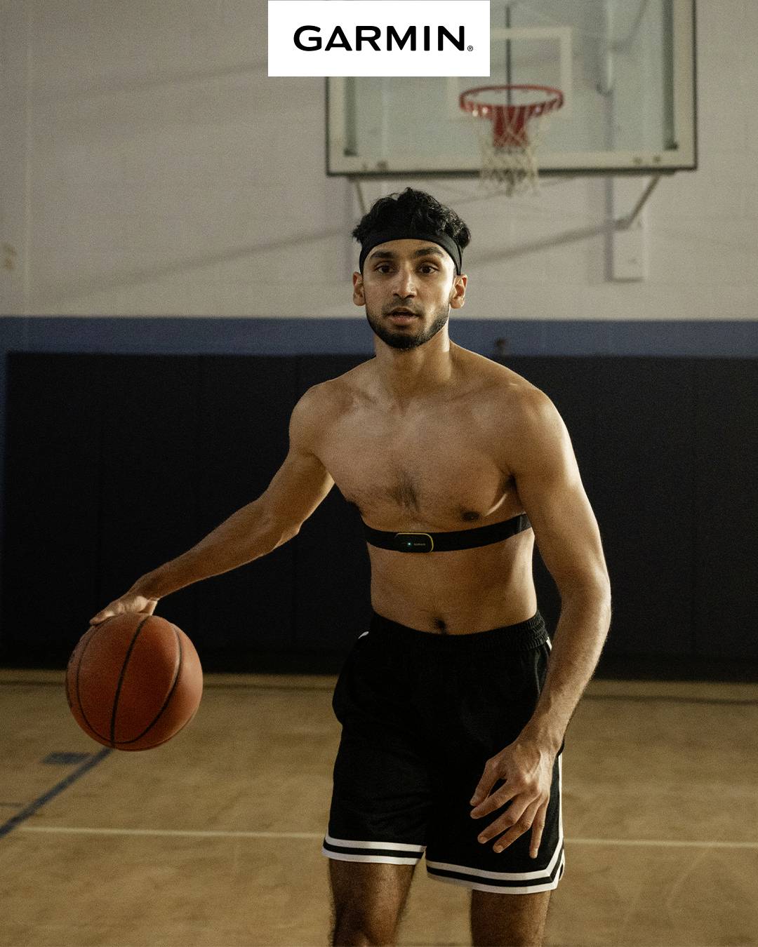 Un homme avec un ballon de basket porte une ceinture cardio de fitness dans un gymnase. En arrière-plan, on peut voir un panier de basket. En haut figure 'GARMIN'.