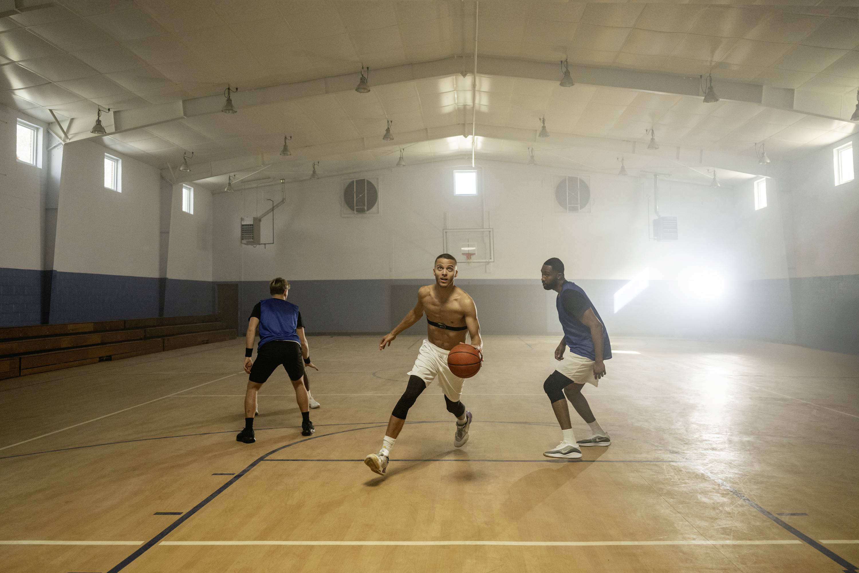 Trois hommes jouent au basketball dans un gymnase. Un joueur dribble le ballon, tandis que les deux autres le défendent.