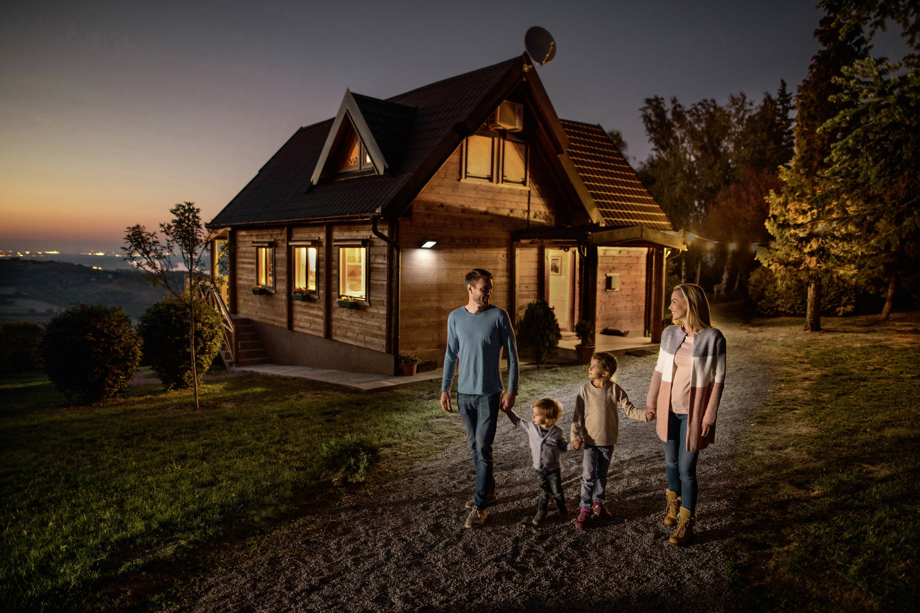 Une famille se promène au coucher du soleil sur un chemin menant à une maison en bois. L'environnement est verdoyant et boisé.