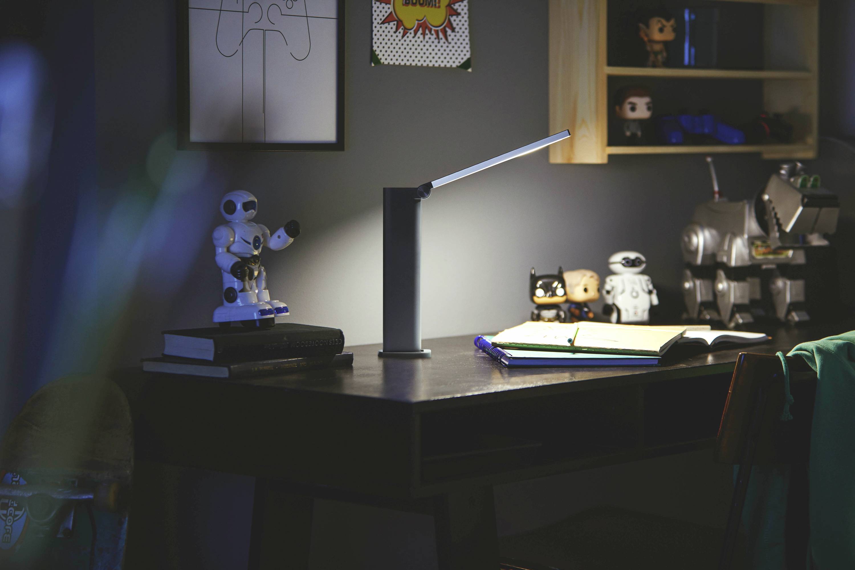 Un bureau avec une lampe allumée éclaire un carnet ouvert. Des robots jouets et des figurines sont visibles en arrière-plan.