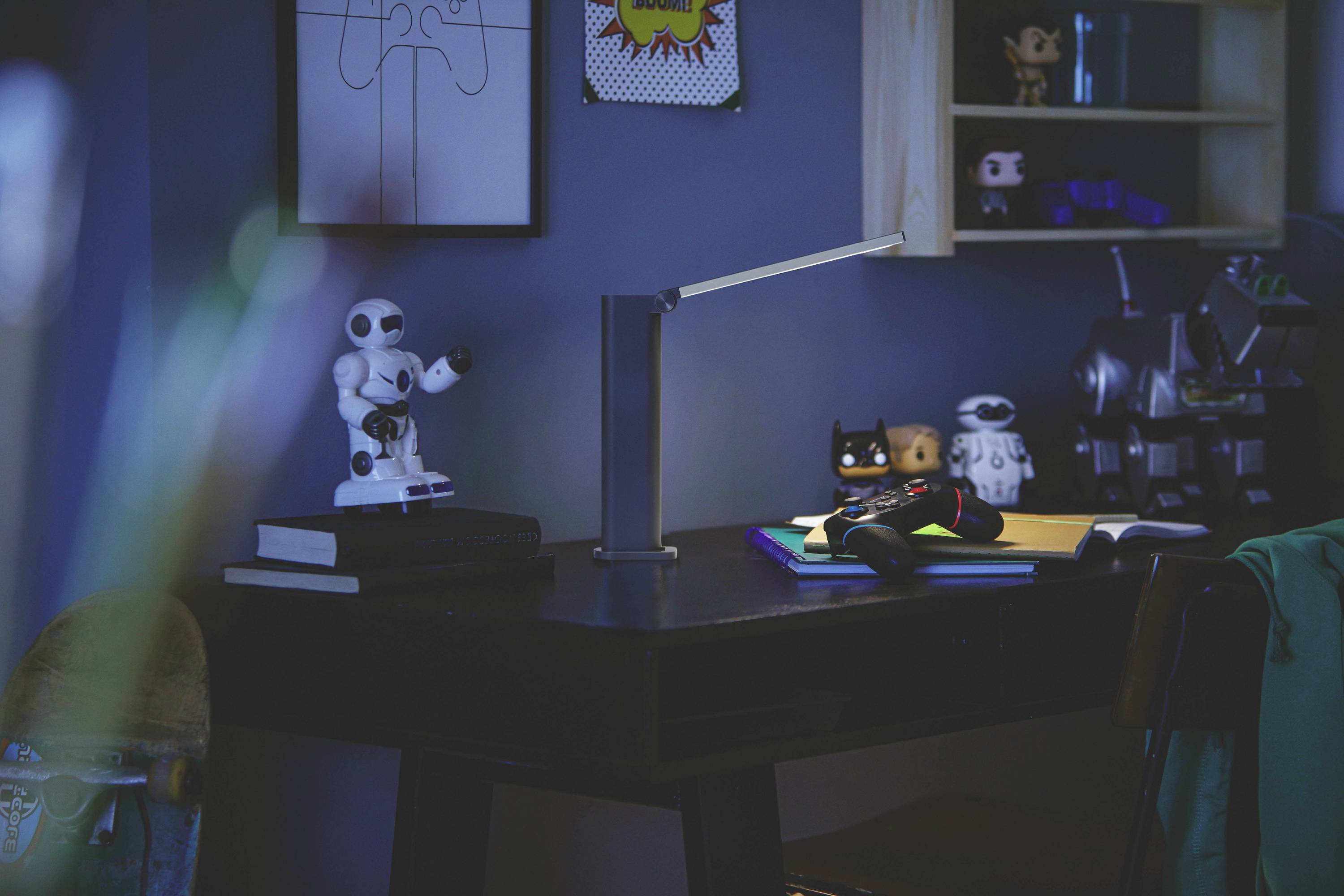 Un bureau avec des figurines et des livres, éclairé par une lampe de bureau. Des robots et des personnages de bande dessinée se trouvent sur l'étagère.
