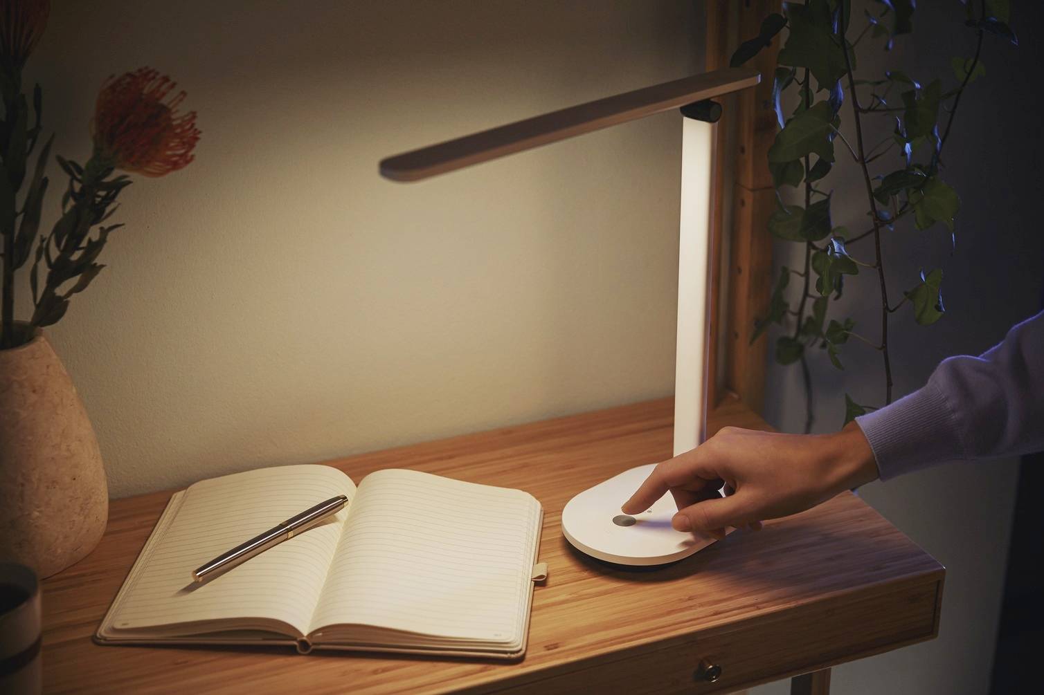 Une personne allume une lampe de bureau moderne. Sur la table se trouvent un carnet ouvert et un stylo.
