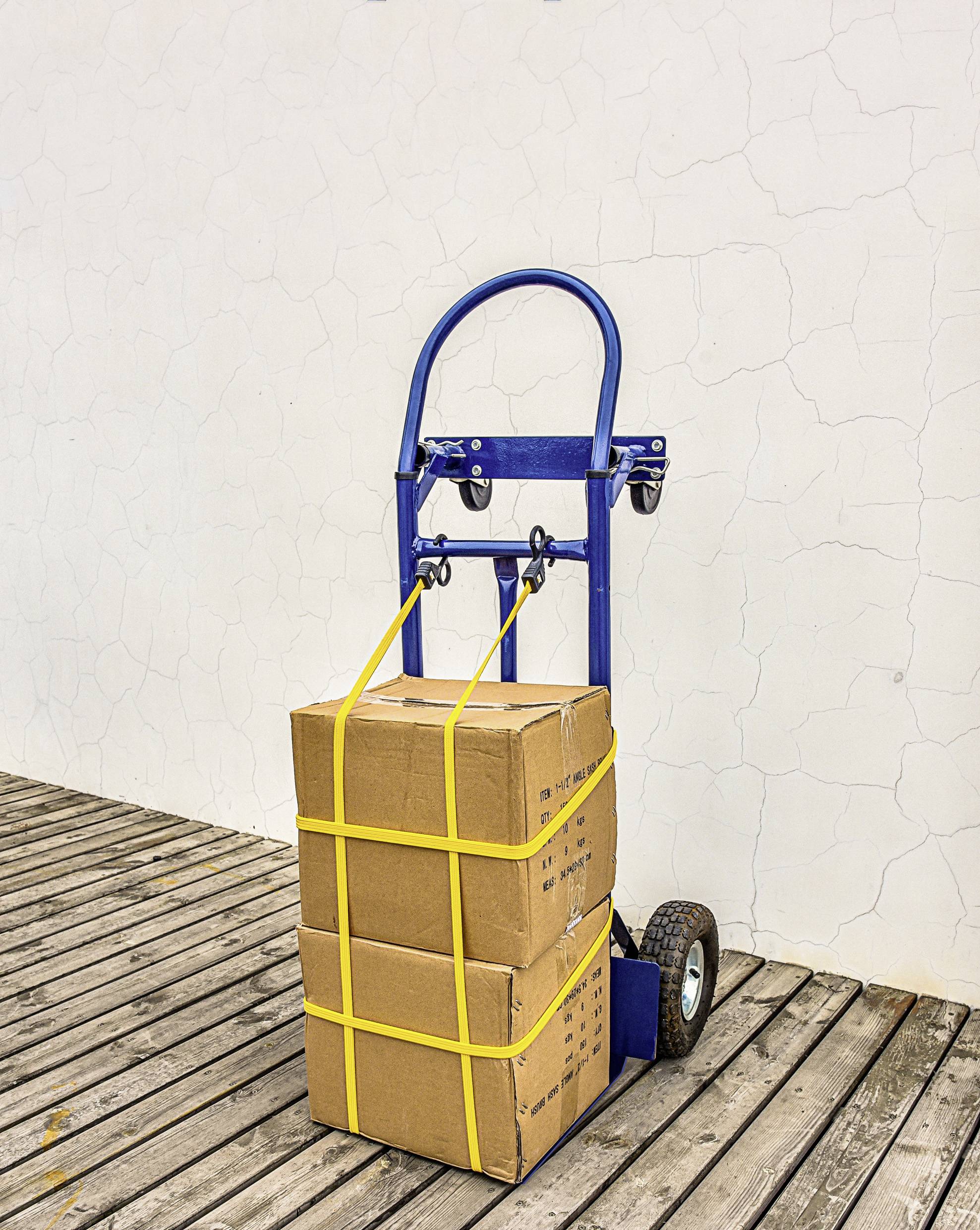 Un chariot à main bleu sur une terrasse en bois transporte deux boîtes en carton empilées, maintenues par des sangles jaunes, posées contre un mur blanc à texture rugueuse.