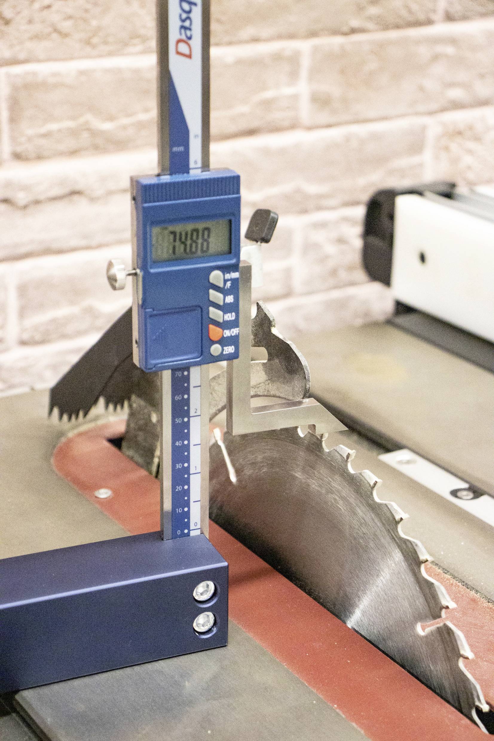 Un pied à coulisse numérique mesure la hauteur d'une lame de scie circulaire sur une scie à table. L'appareil affiche '74,08', indiquant une mesure de précision.