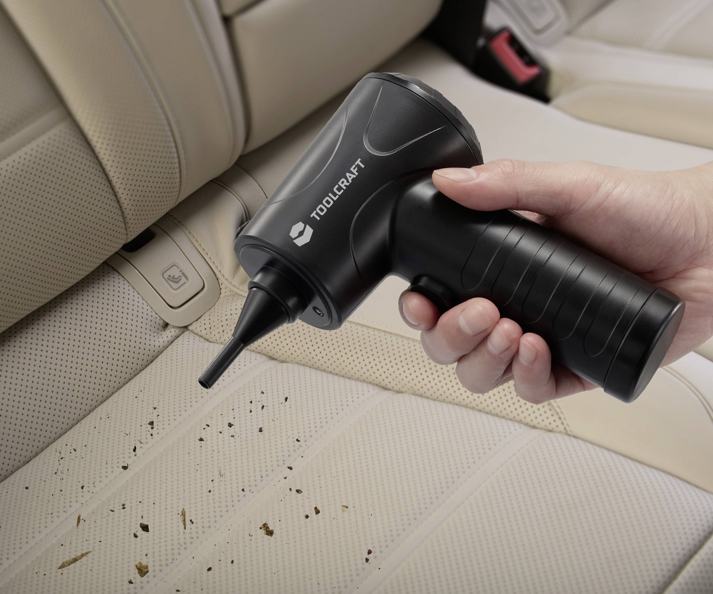Une personne utilise un aspirateur portable noir pour nettoyer des miettes sur un siège de voiture beige. L'aspirateur est de la marque 'Toolcraft'.