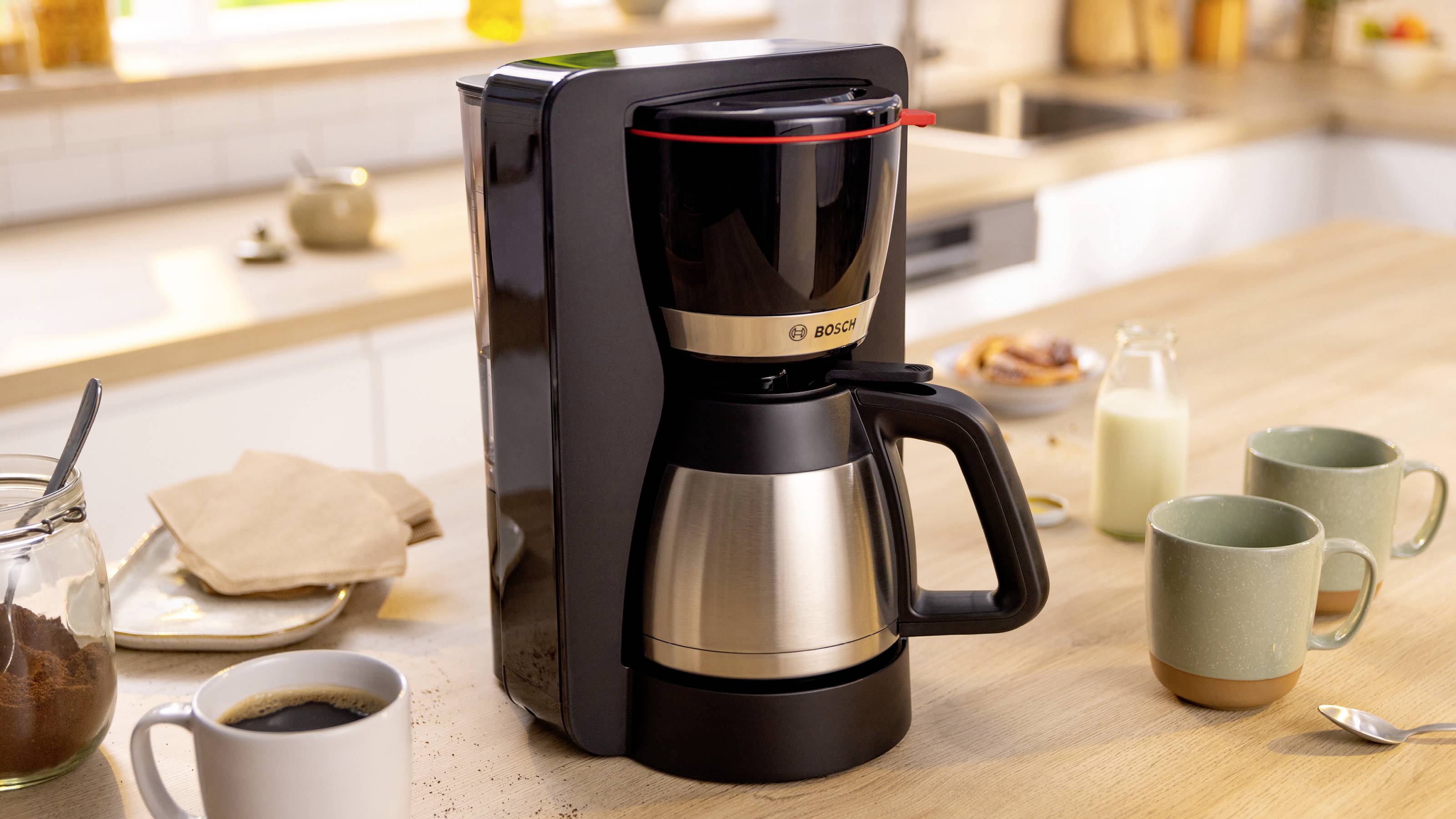 Une machine à café sur un comptoir de cuisine, entourée de mugs, d'un bocal de café, d'un pot de lait et d'un sucrier, suggérant la préparation du café du matin.