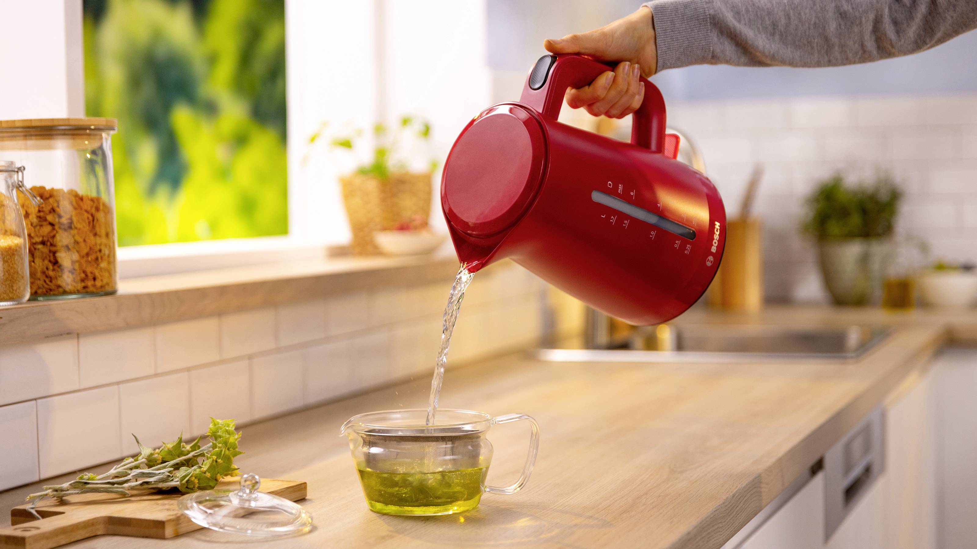 Une personne verse de l'eau chaude d'une bouilloire électrique rouge dans une tasse en verre avec un infuseur à thé sur un plan de travail de cuisine, près d'une fenêtre.