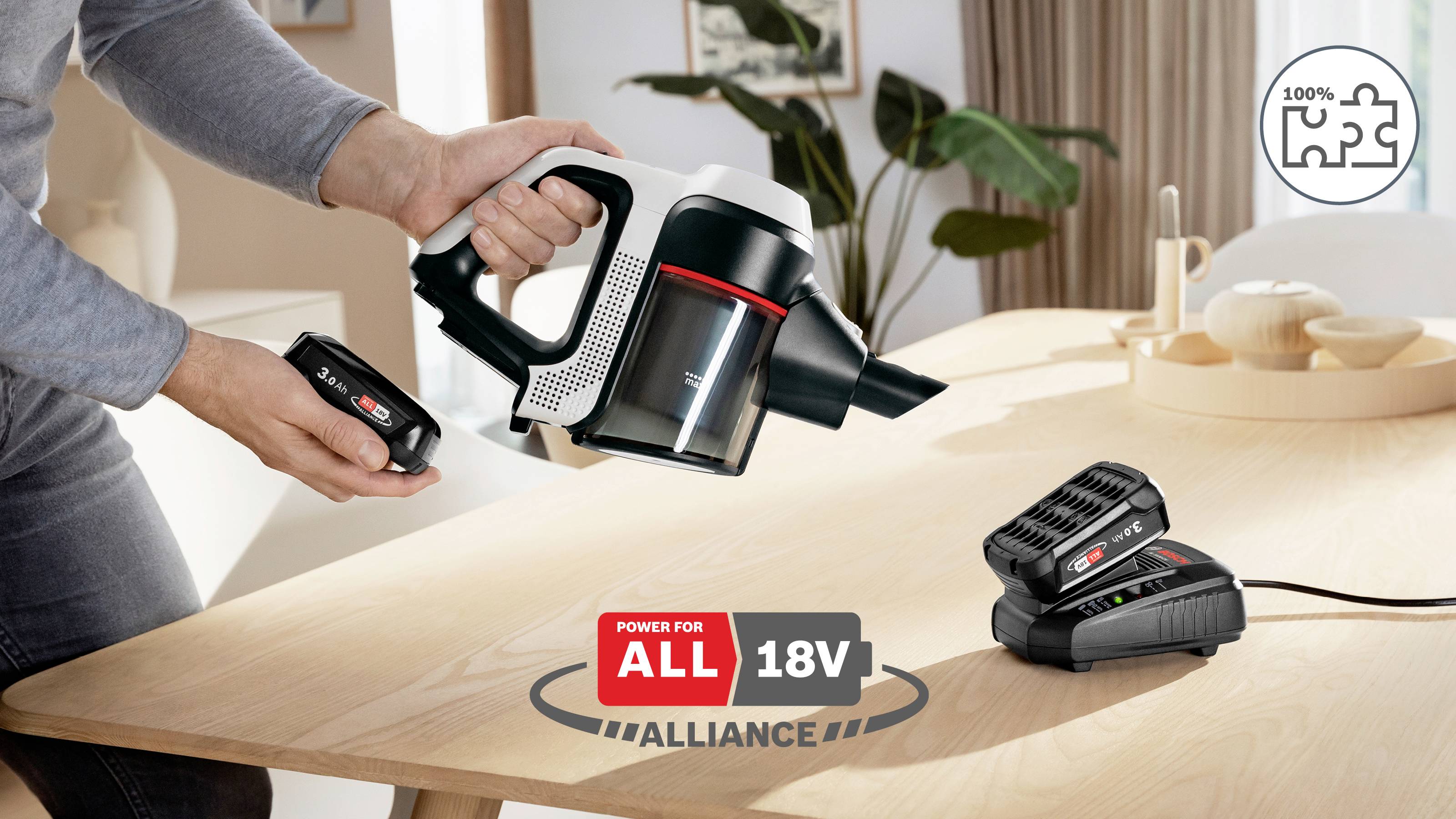 Alliance 18V « Pouvoir pour tous » : aspirateur présenté avec une batterie amovible, un chargeur et une icône indiquant une compatibilité à 100 % sur une table en bois.