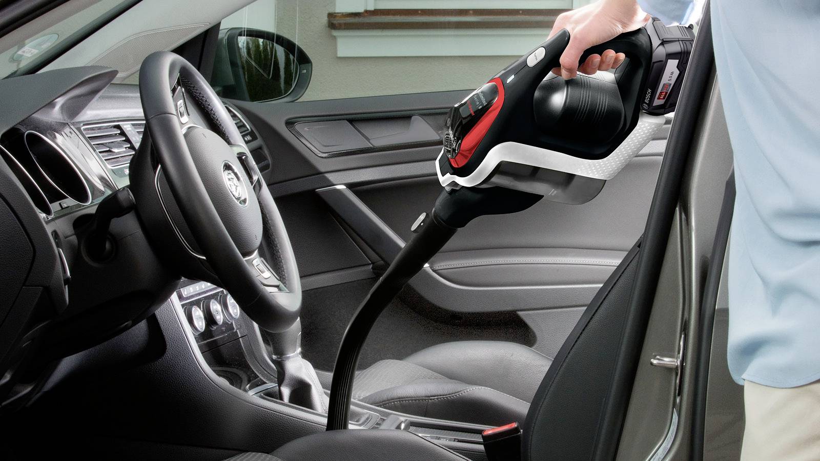 Une personne aspirant l'intérieur d'une voiture avec un aspirateur portable, concentrée sur le siège avant du passager et la zone de la console centrale.