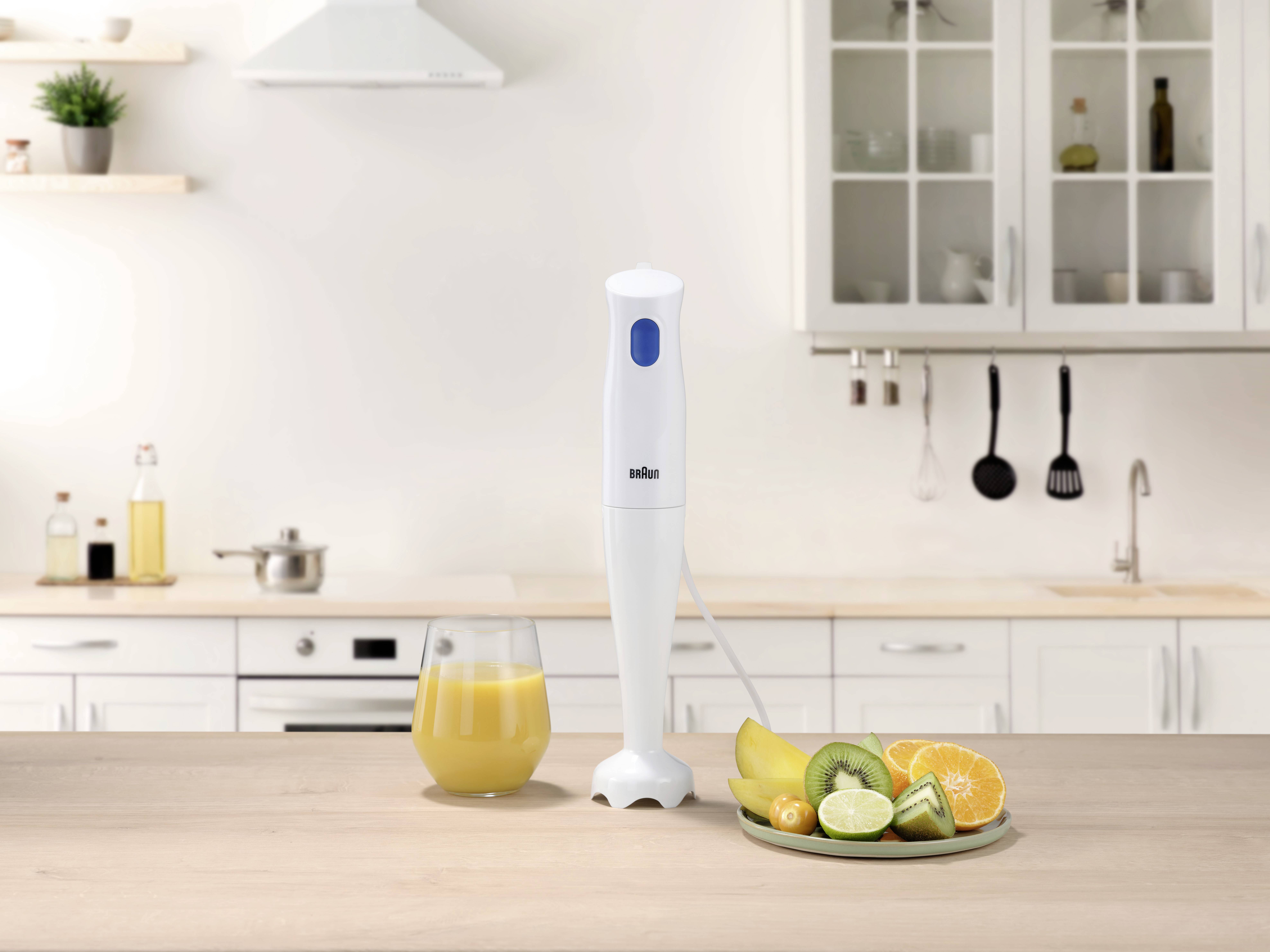 Un mixeur plongeant blanc est posé sur un plan de travail de cuisine, à côté d'un verre de jus jaune et d'une assiette de fruits tranchés comprenant des oranges et des kiwis.