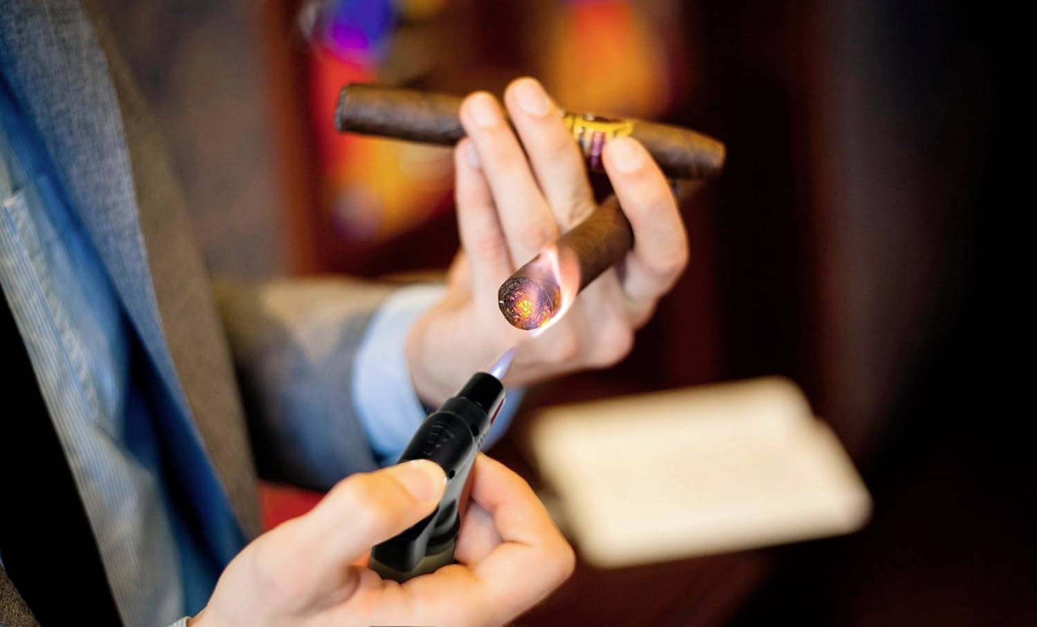 Un homme en costume allume un cigare avec un briquet de table, se concentrant sur l'extrémité incandescente, dans une pièce doucement éclairée.