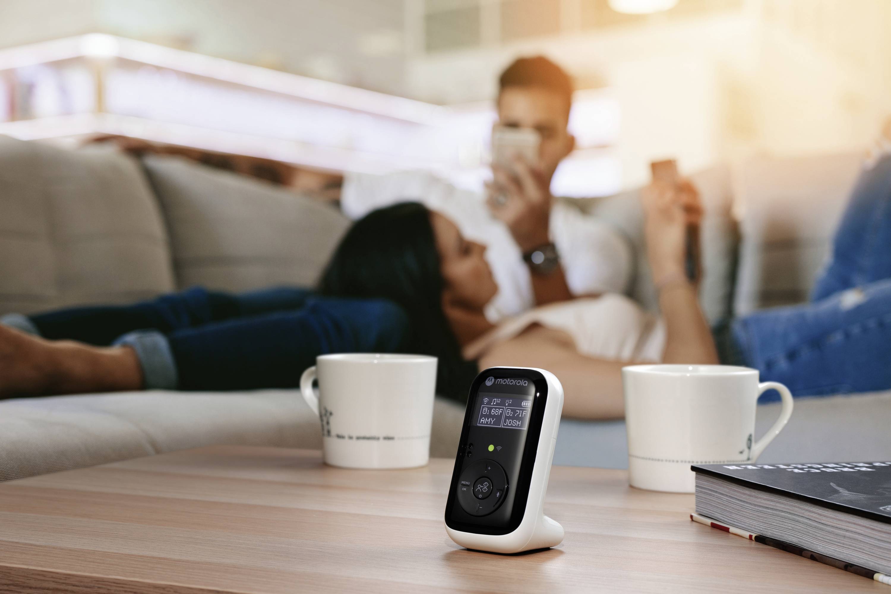 Un baby-phone est posé sur une table au premier plan, à côté de deux tasses à café, tandis qu'un couple se détend sur un canapé en arrière-plan, dans un flou artistique.