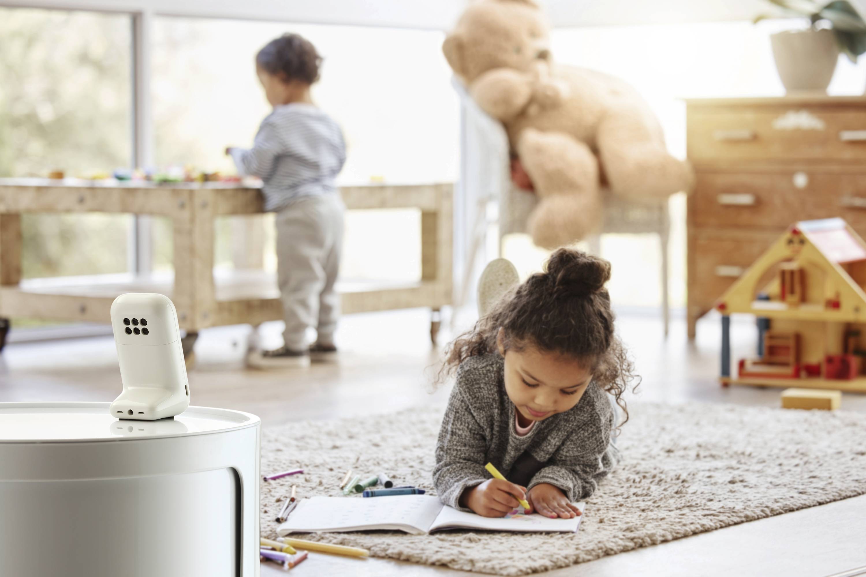 Un enfant dessine sur du papier en étant allongé sur un tapis. Un autre enfant joue à une table en arrière-plan. Un babyphone est visible à proximité.