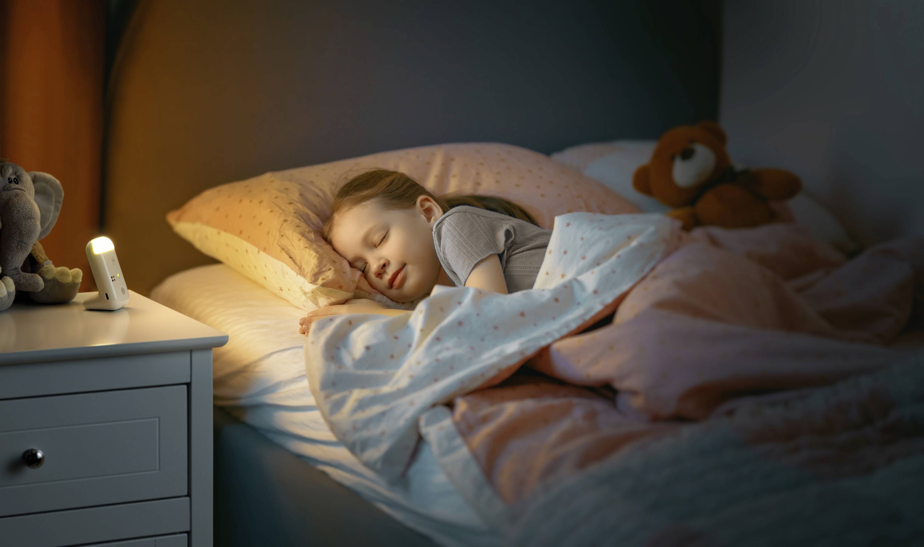 Un enfant dort paisiblement dans son lit, avec une petite veilleuse posée sur la table de chevet. Un ours en peluche est installé à côté de l'oreiller.