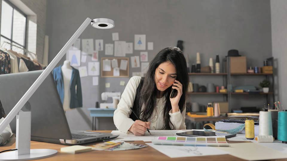 Une femme dans un bureau à domicile fait du multitâche en parlant au téléphone tout en écrivant à un bureau avec un ordinateur portable, des nuanciers de couleurs et du matériel de couture.