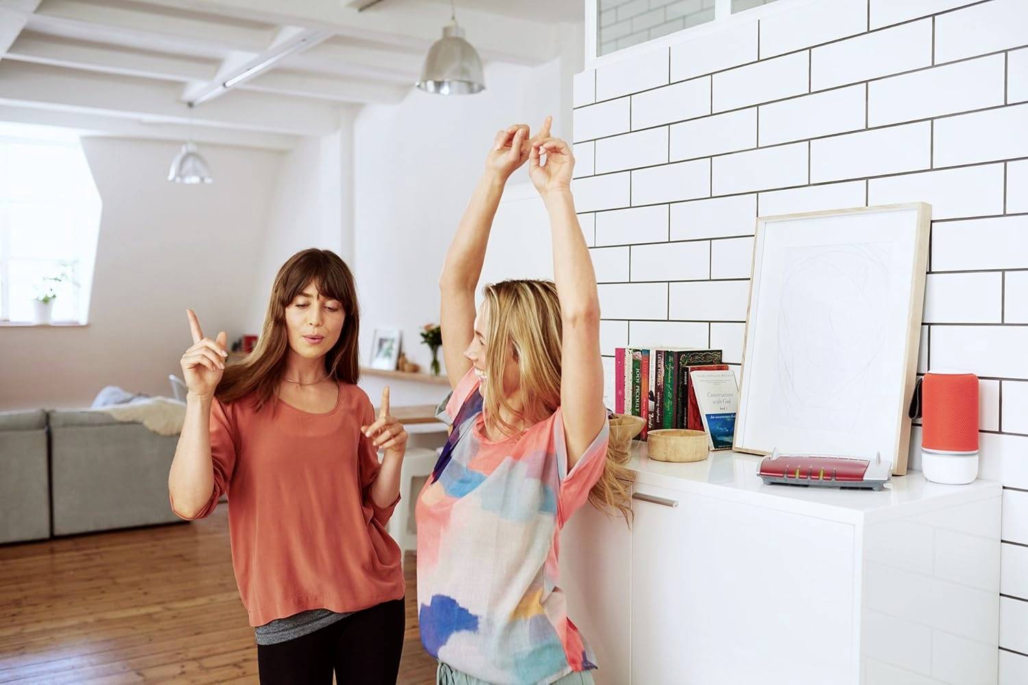 Deux femmes dansant dans un salon lumineux avec des planchers en bois et des murs en briques blanches, entourées de livres et de décoration moderne.