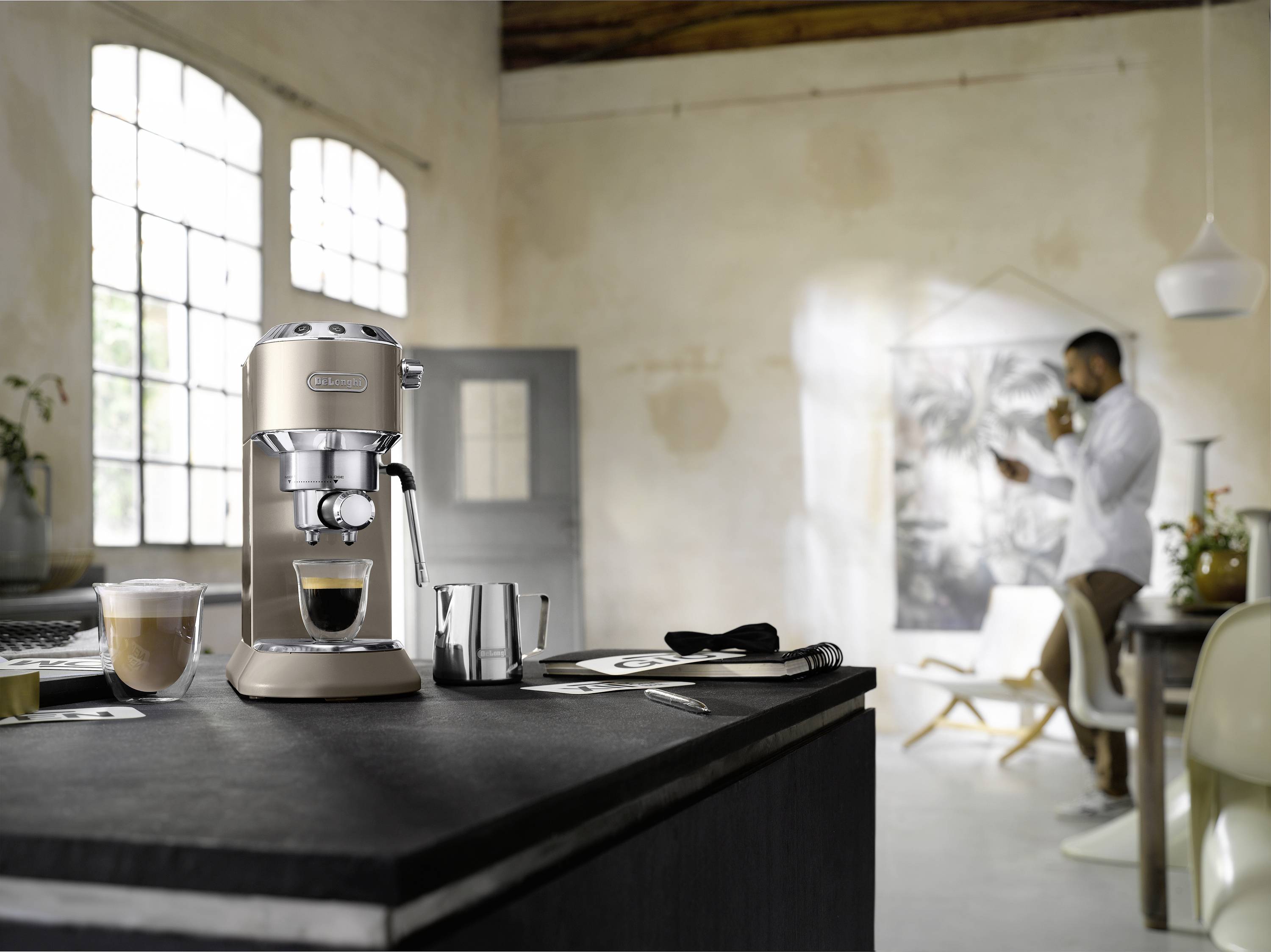 Une machine à espresso moderne sur un comptoir de cuisine avec deux verres de café ; une personne se tient en arrière-plan en sirotant une boisson.