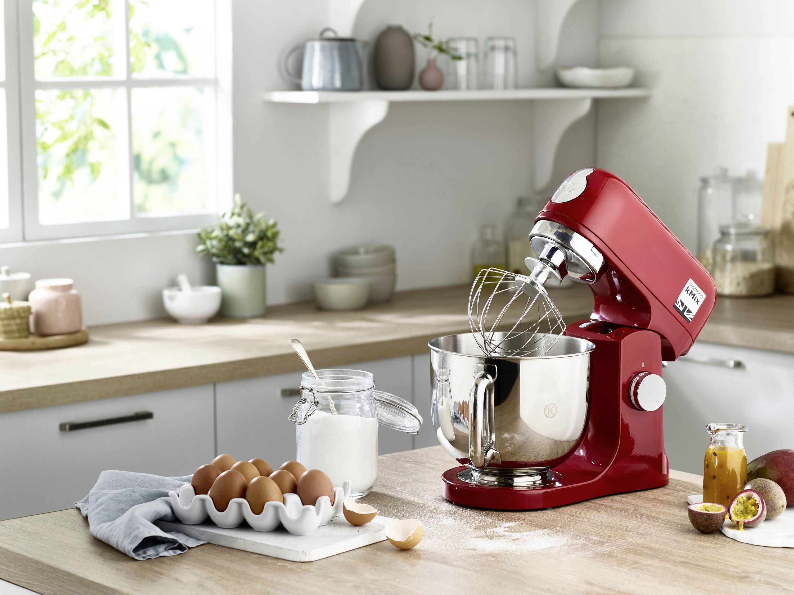 Un batteur mélangeur rouge sur un plan de travail de cuisine avec un fouet attaché dans un bol en métal. À proximité se trouvent un pot de sucre et une boîte d'œufs.