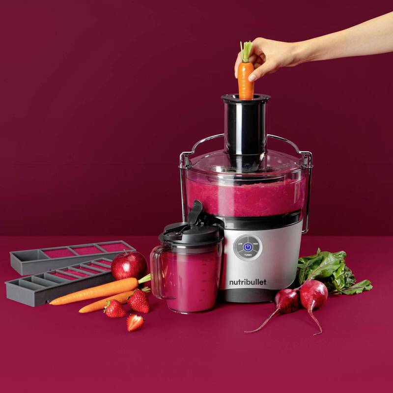 Une personne insère une carotte dans un extracteur de jus Nutribullet, entourée de fraises, de carottes et de betteraves, avec un jus fini devant.