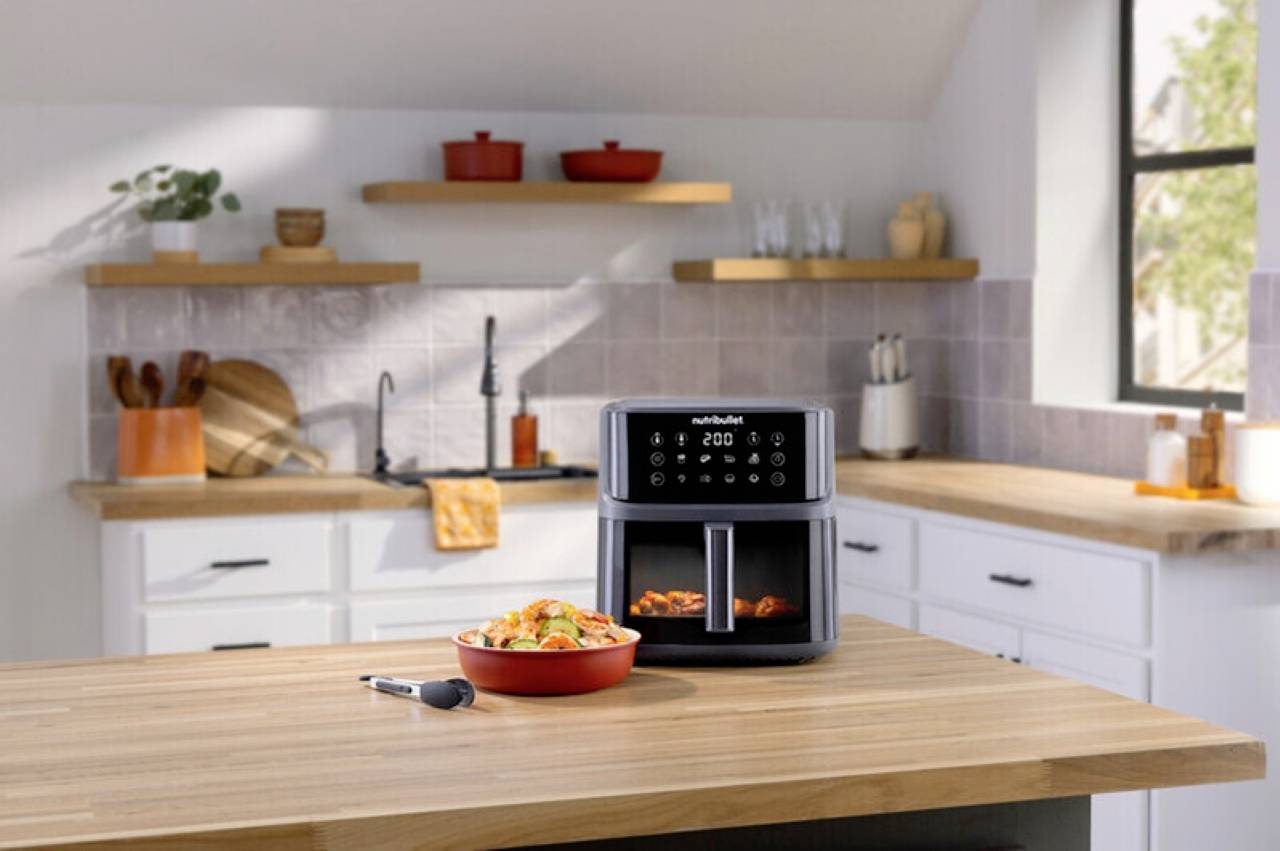 Un air fryer sur un comptoir de cuisine avec du poulet cuit et des légumes dans un bol à côté. Des étagères de cuisine et un évier en arrière-plan.