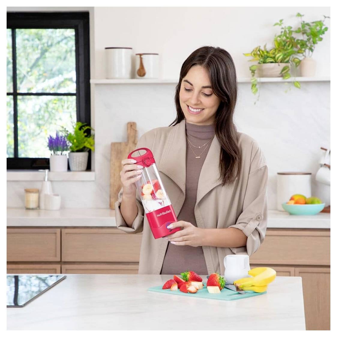 Une personne dans une cuisine tenant un petit blender rempli de fruits, avec des morceaux de fruits et une plante visibles en arrière-plan.