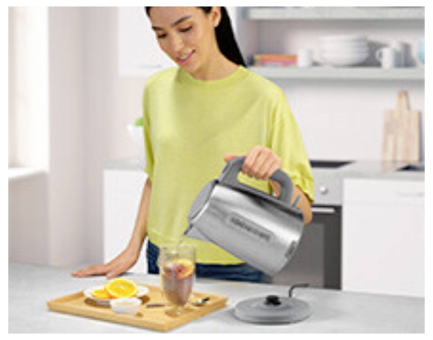 Une personne dans une cuisine verse de l'eau chaude d'une bouilloire électrique dans un verre de thé. Une planche à découper avec des oranges tranchées et des sachets de thé se trouve à proximité.