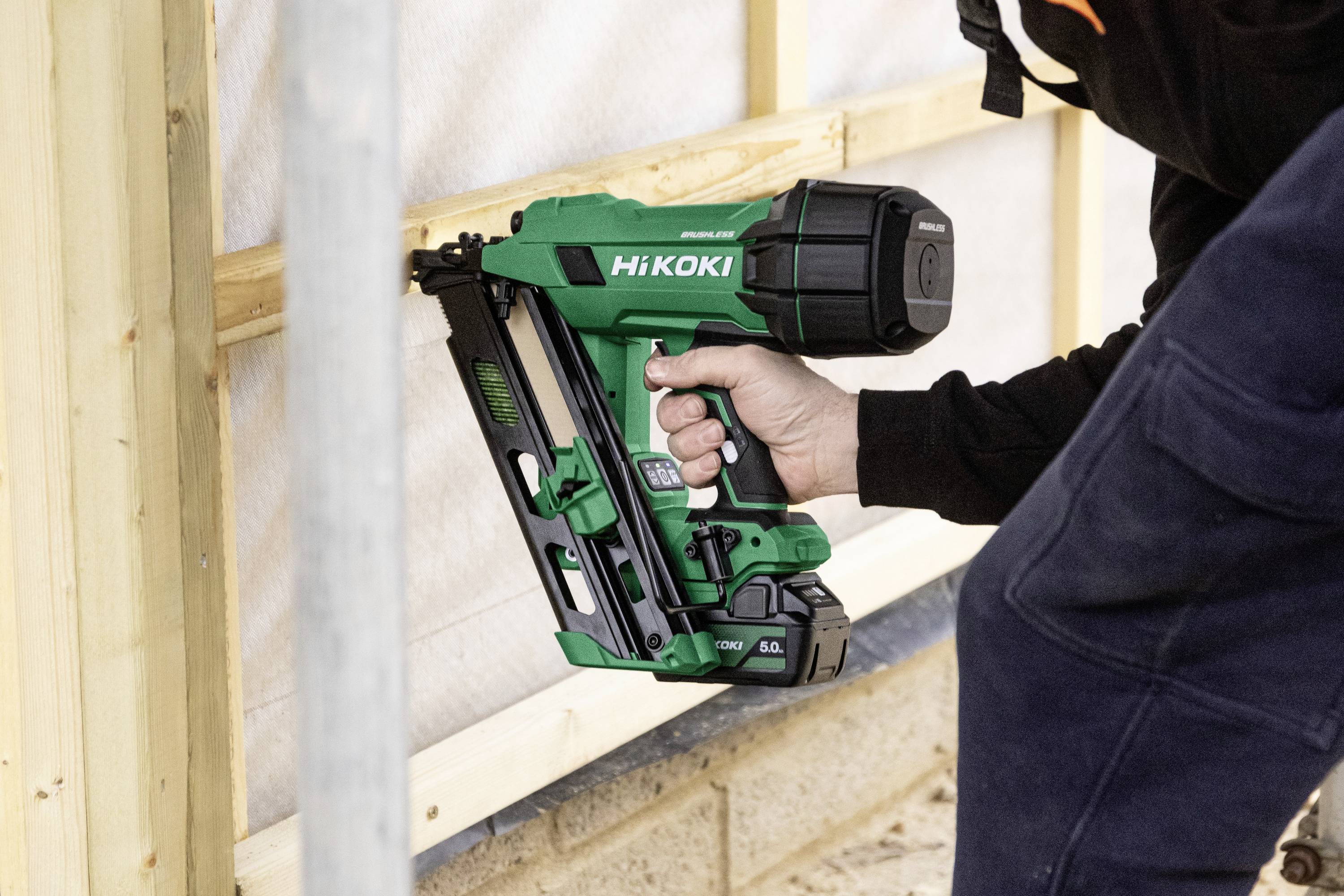 Une personne utilise un cloueur HiKOKI vert pour fixer des cadres en bois sur un chantier de construction extérieur.