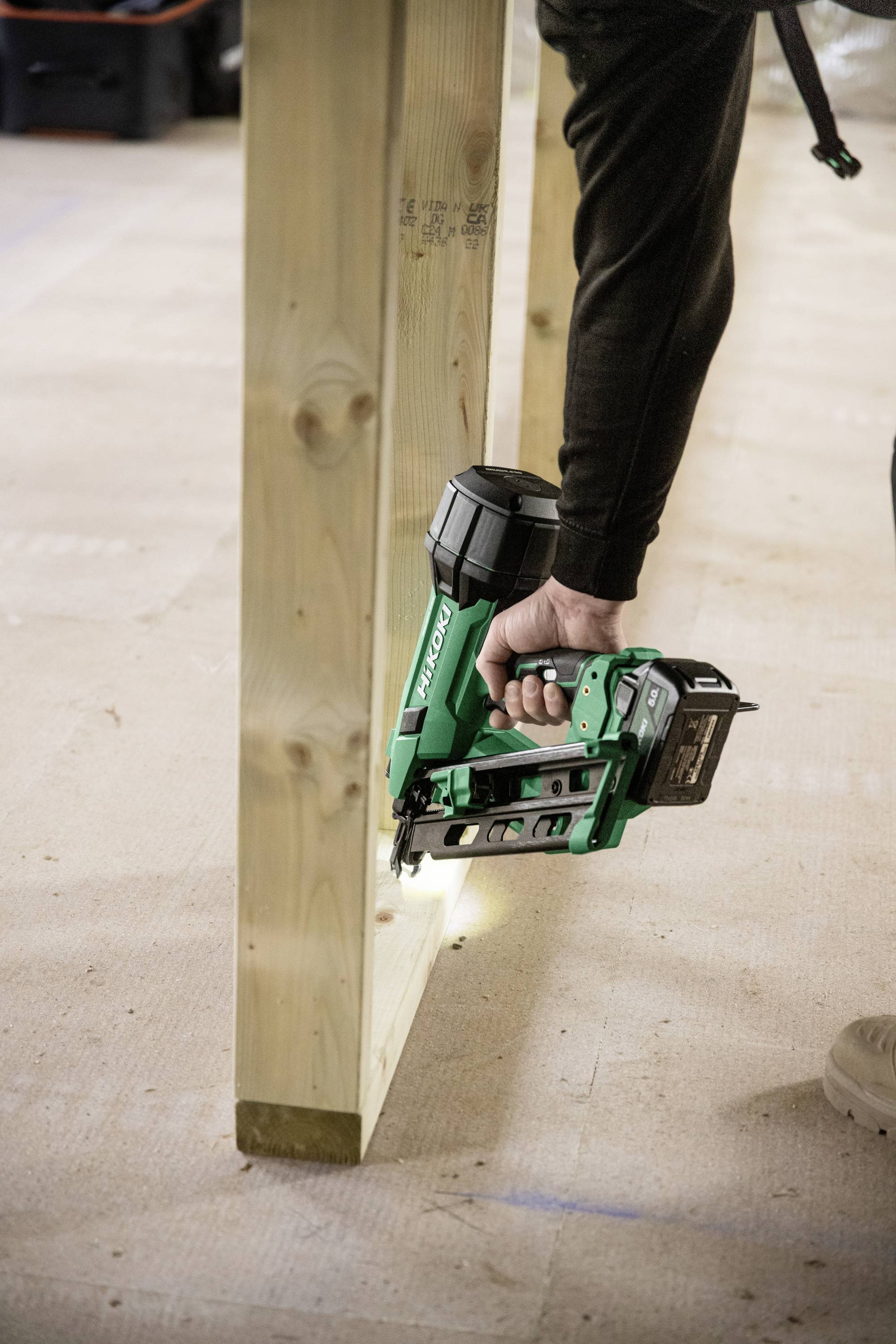 Une personne utilise un pistolet de clouage sans fil vert pour assembler deux poutres en bois sur un chantier de construction.