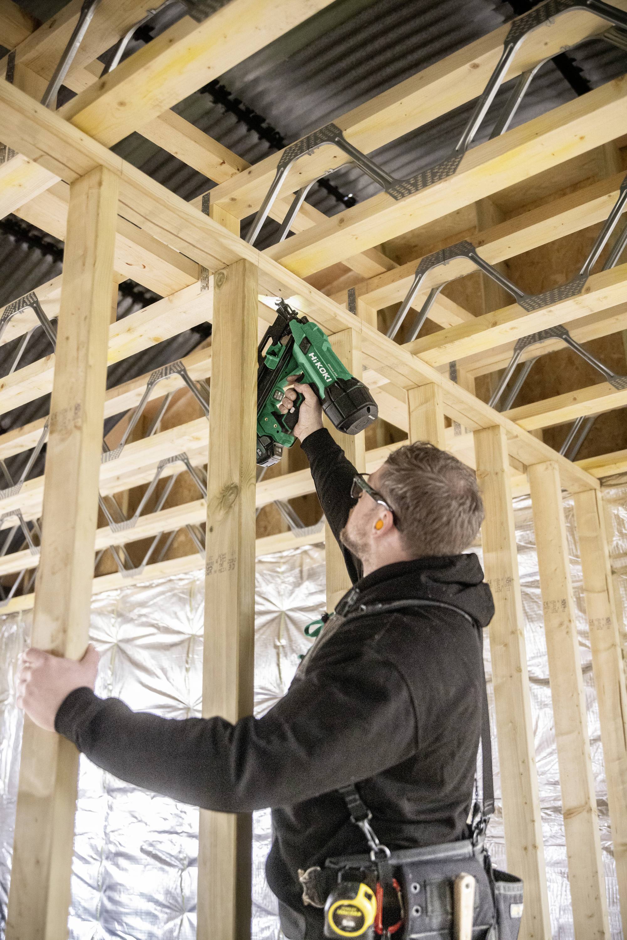 Un ouvrier du bâtiment utilise un cloueur pneumatique pour fixer l'ossature en bois à l'intérieur d'un bâtiment en cours de construction. L'isolation et le câblage sont visibles.