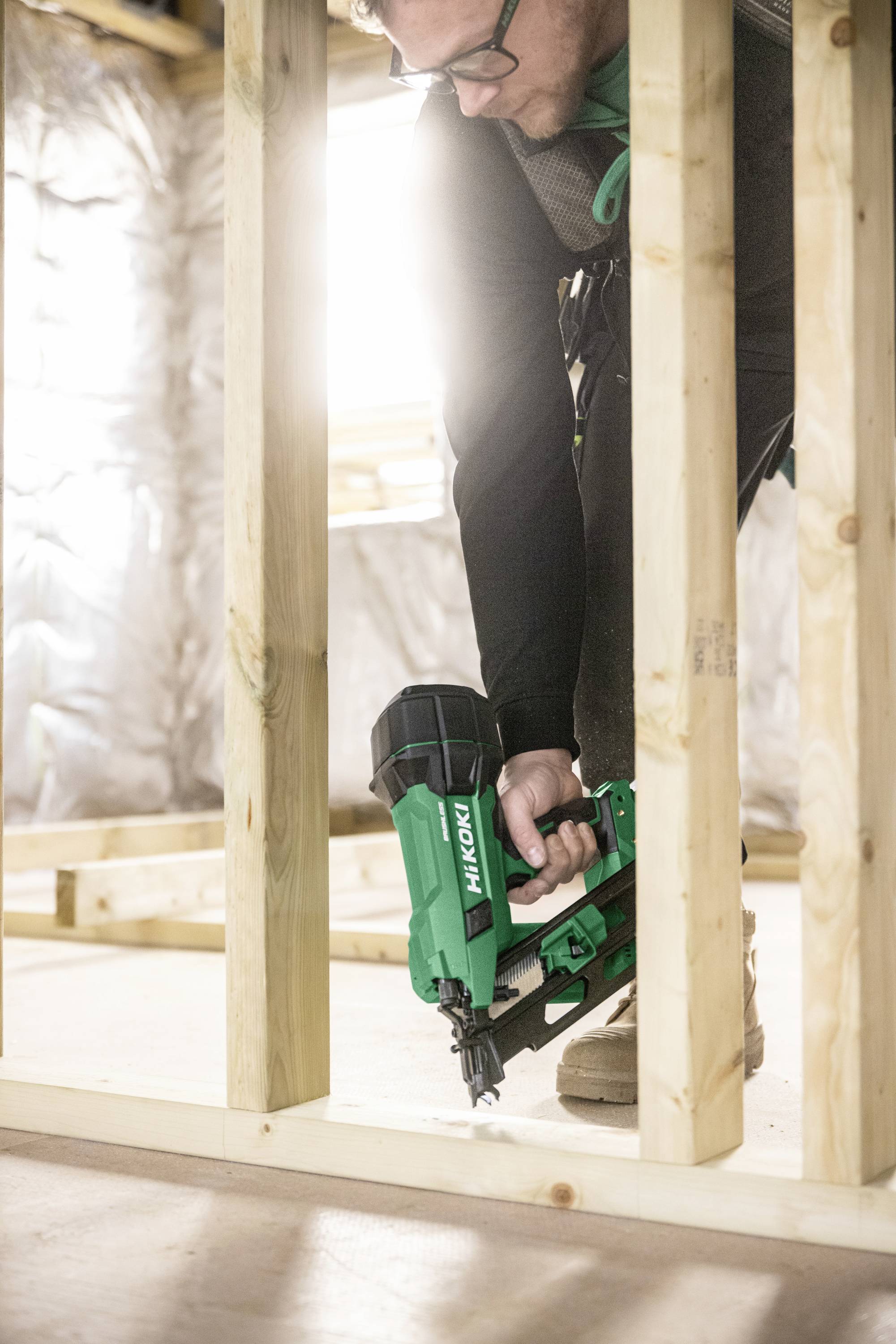 Une personne utilise un pistolet de clouage vert pour fixer des cadres en bois sur un chantier de construction. La lumière du soleil filtre à travers, illuminant l'espace de travail.