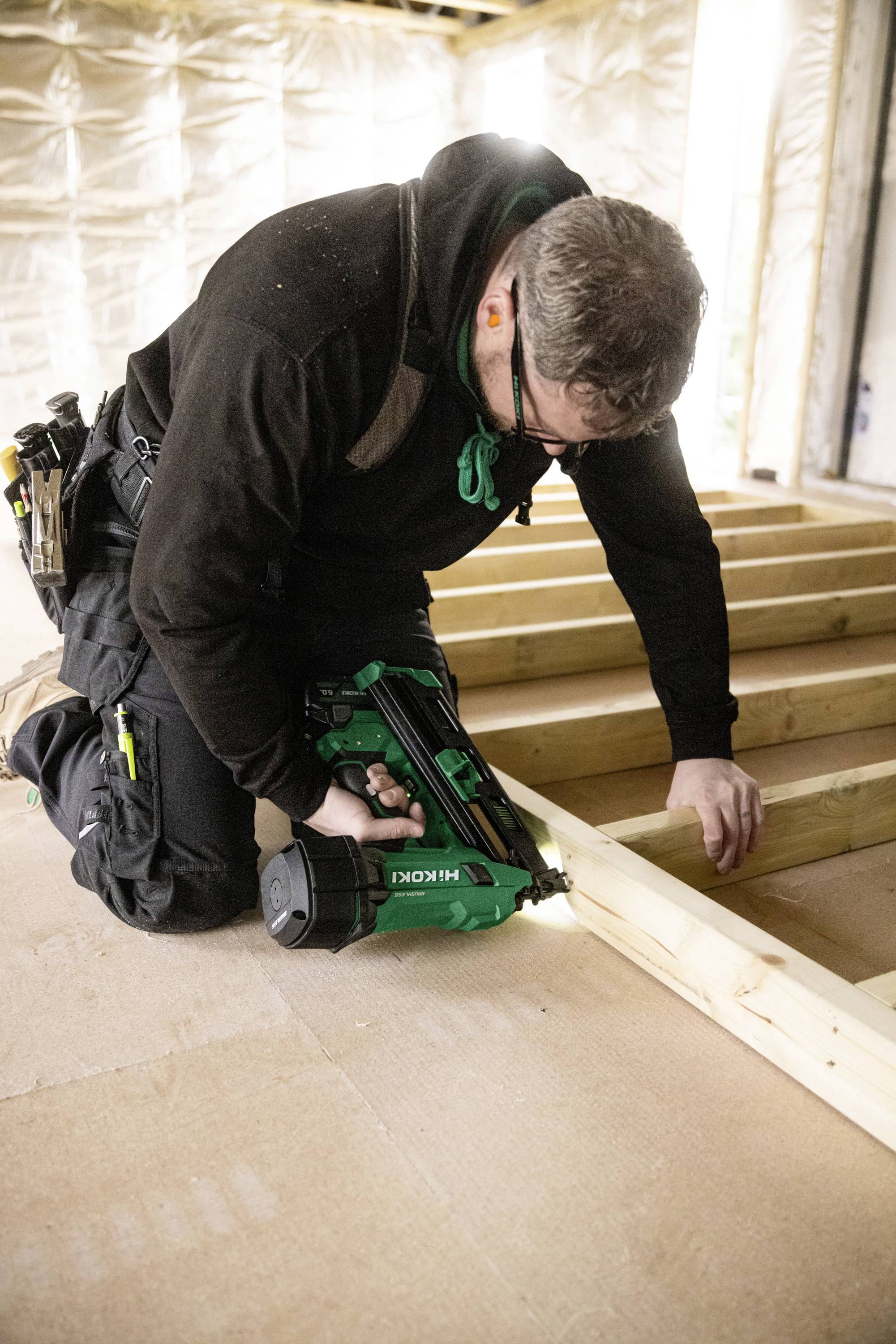 Une personne est à genoux sur un plancher en bois, utilisant un pistolet à clous vert pour assembler des poutres en bois, suggérant un travail de construction ou de charpenterie en cours.