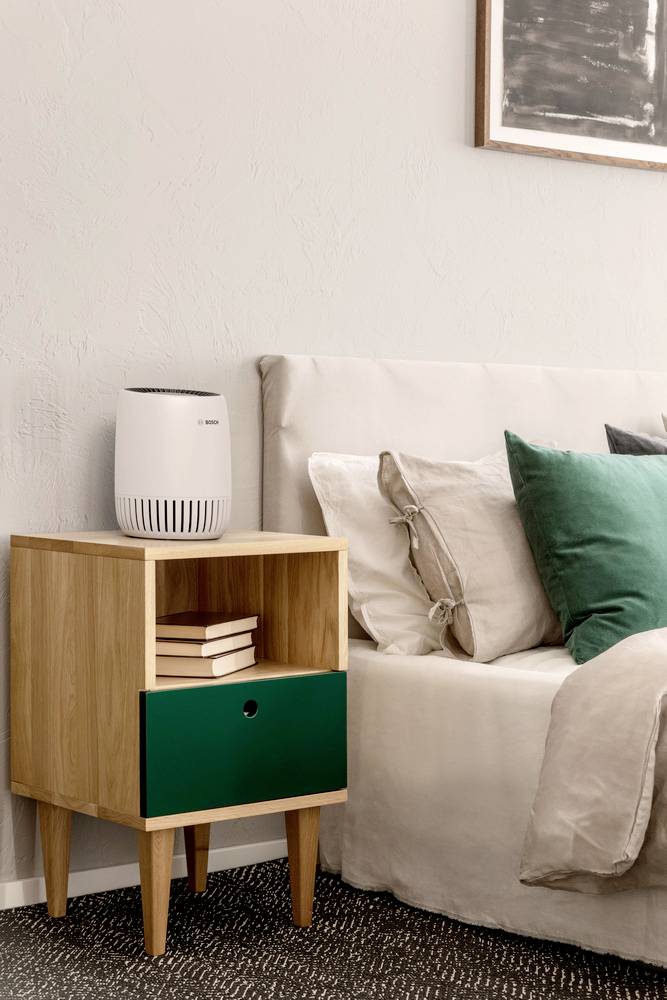 Une chambre soigneusement rangée avec une table de chevet en bois sur laquelle sont posés des livres, un petit purificateur d'air blanc, à côté d'un lit avec des oreillers verts et beiges.