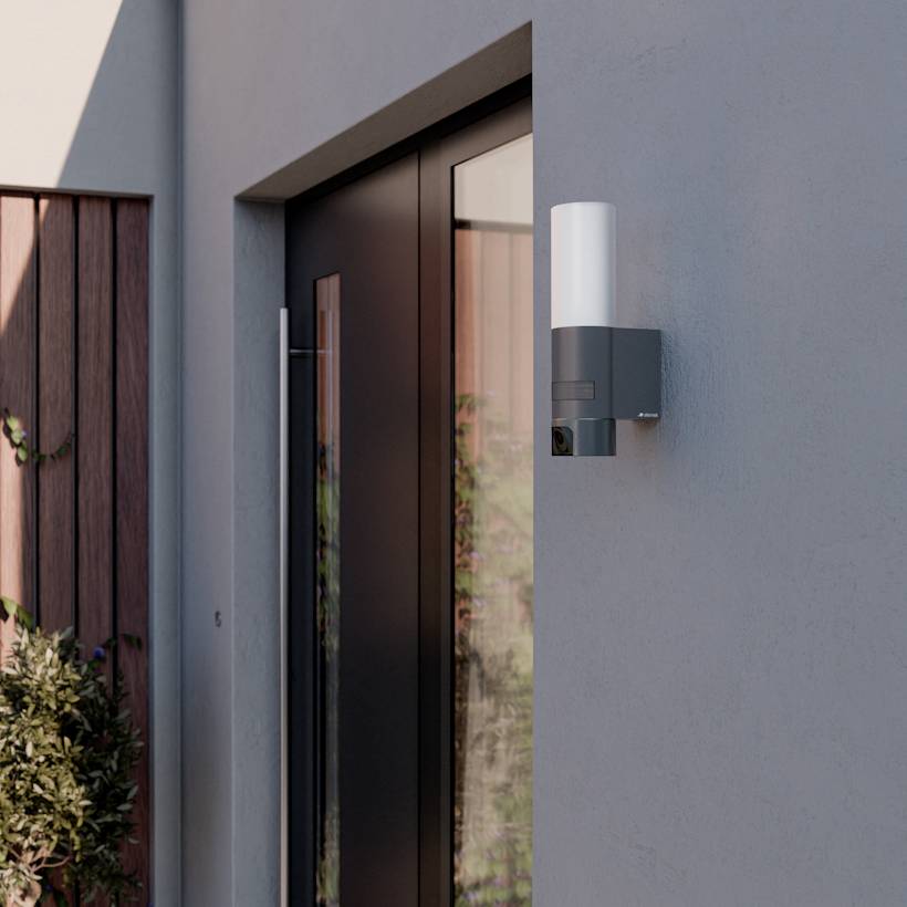 Un éclairage de sécurité extérieur moderne installé à côté d'une porte sur un mur gris, avec une clôture en bois et des arbustes visibles en arrière-plan.