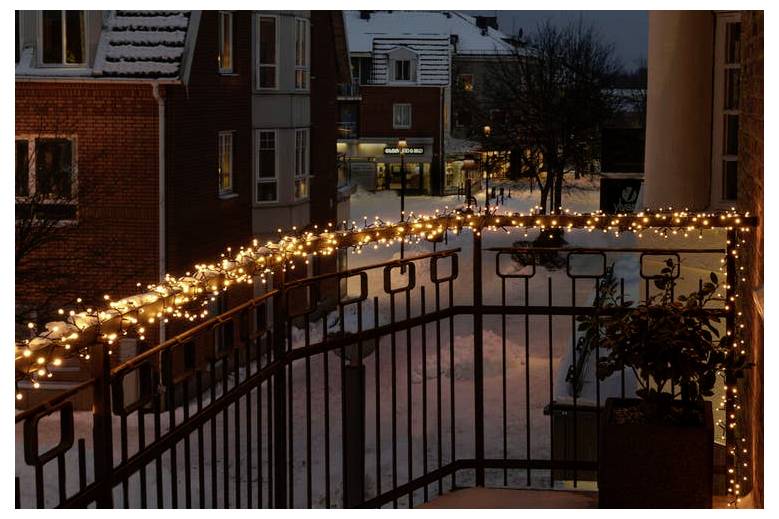 Une rue enneigée vue depuis un balcon décoré de guirlandes lumineuses scintillantes, créant une atmosphère festive dans un quartier paisible au crépuscule.
