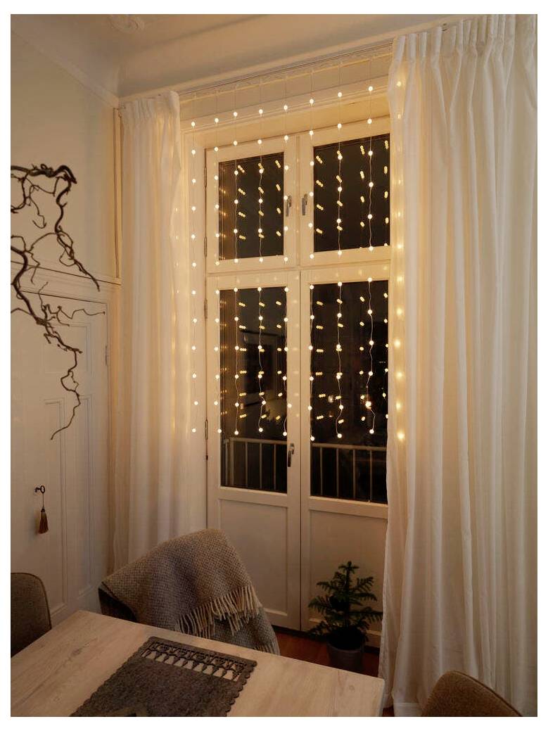 Une fenêtre ornée de guirlandes lumineuses illumine une pièce chaleureuse avec des rideaux blancs, une petite plante verte et une table sur laquelle est posé un plaid plié.