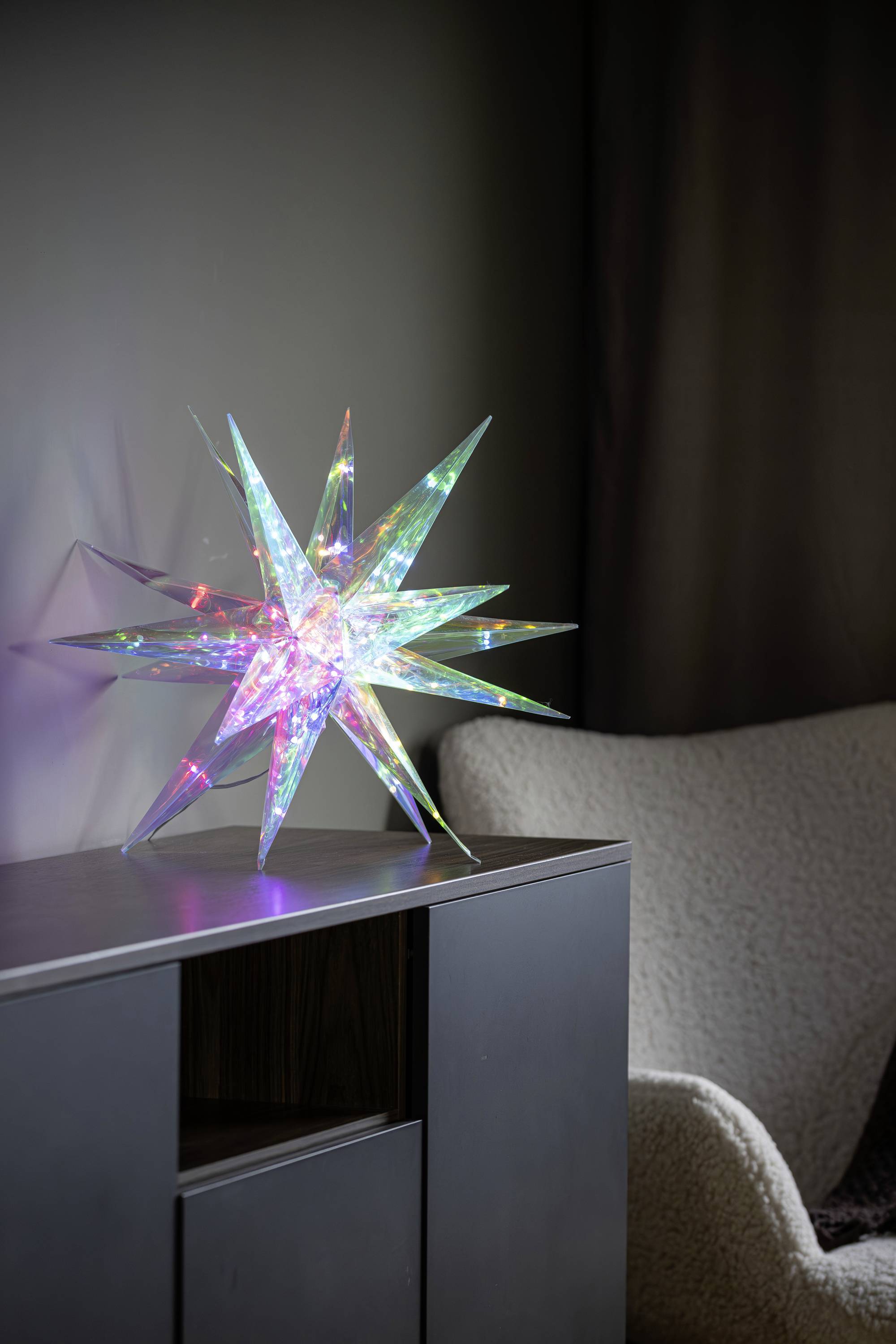 Une sculpture d'étoile lumineuse et colorée aux pointes acérées est posée au sommet d'un meuble sombre dans une pièce faiblement éclairée, près d'une chaise blanche à texture.