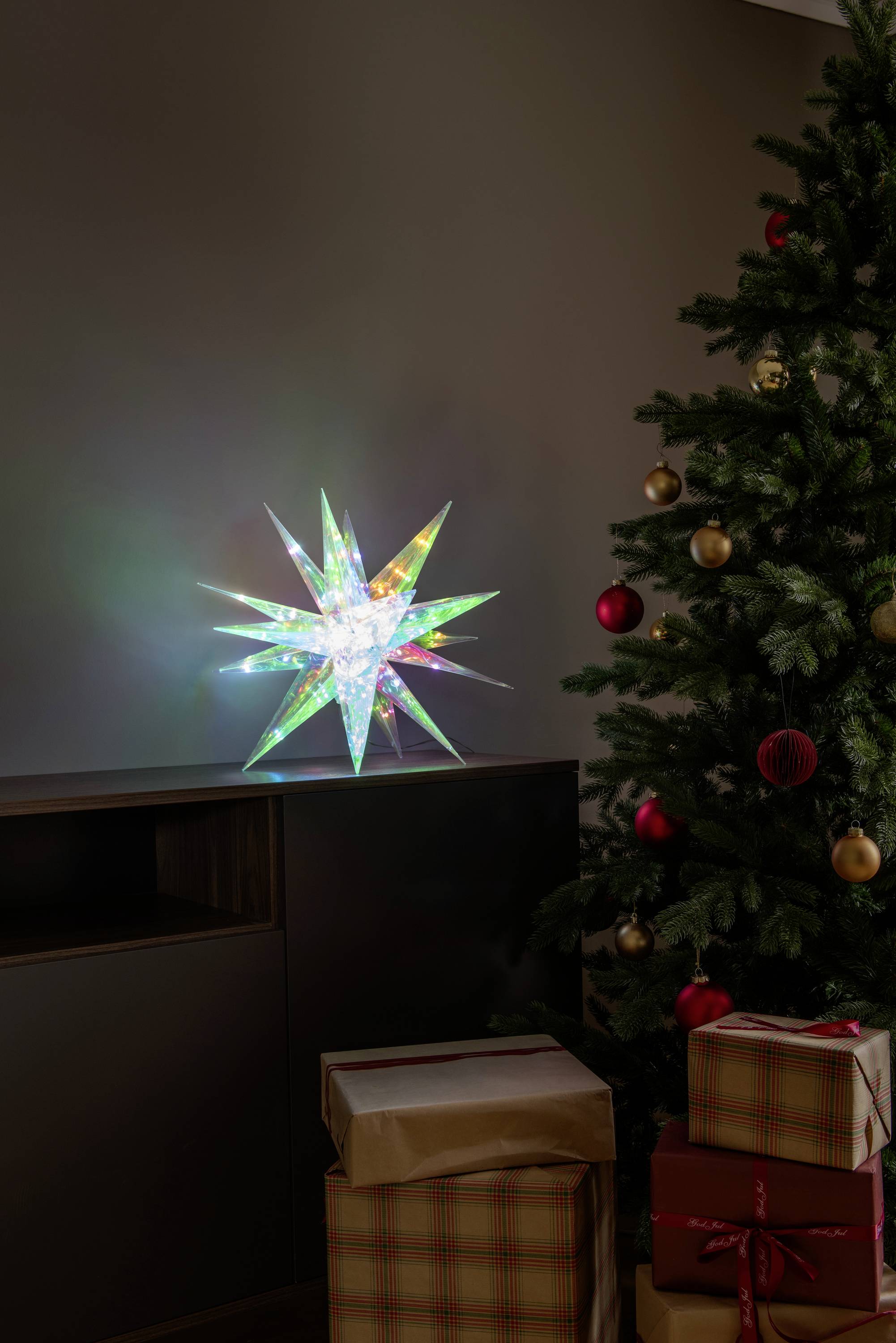 Une décoration d'étoile lumineuse multicolore est posée sur une étagère près d'un arbre de Noël décoré et de cadeaux emballés.