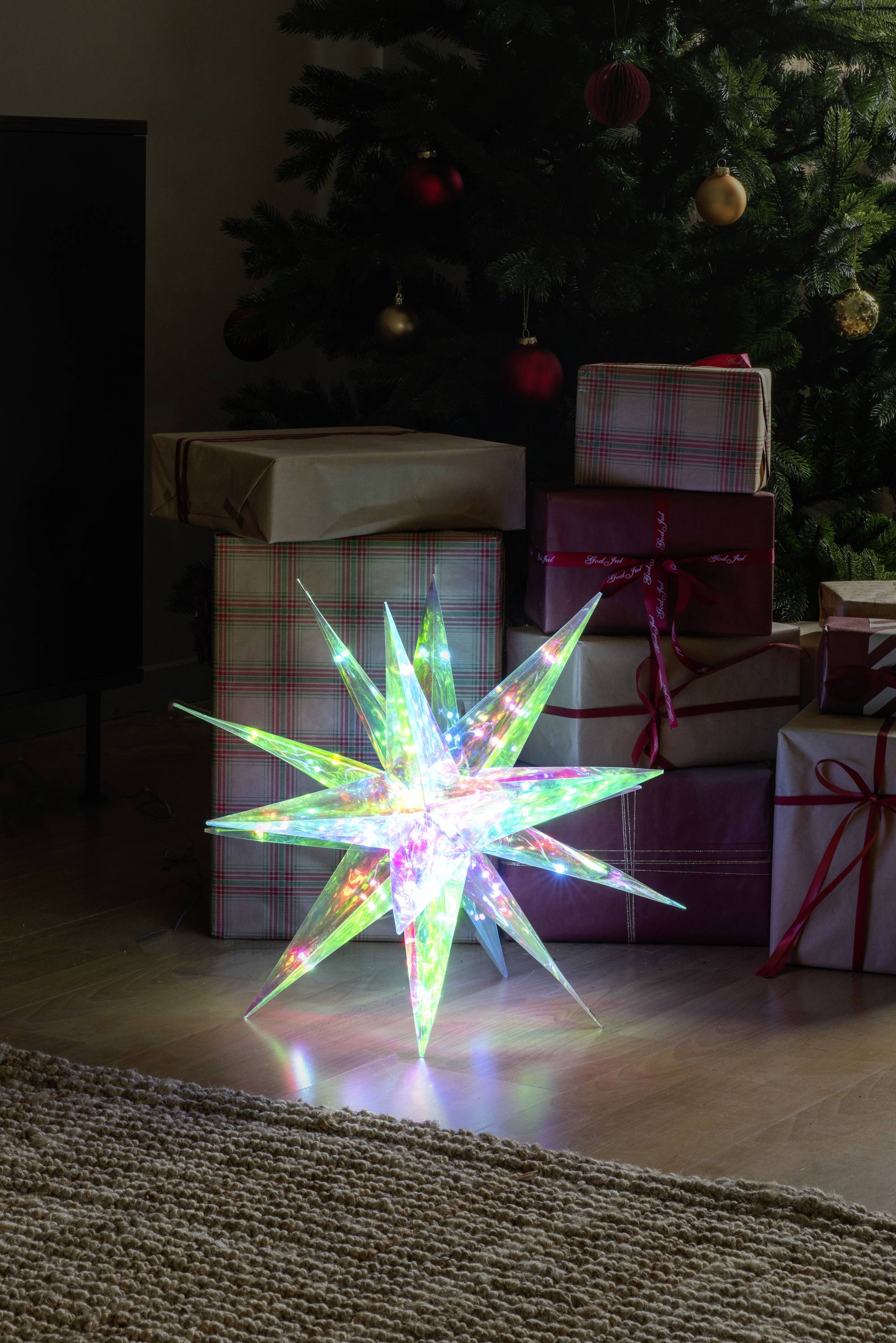 Une lumière étoilée scintillante, multicolore, est posée sur le sol devant des cadeaux de Noël empilés et emballés, à côté d'un arbre décoré.