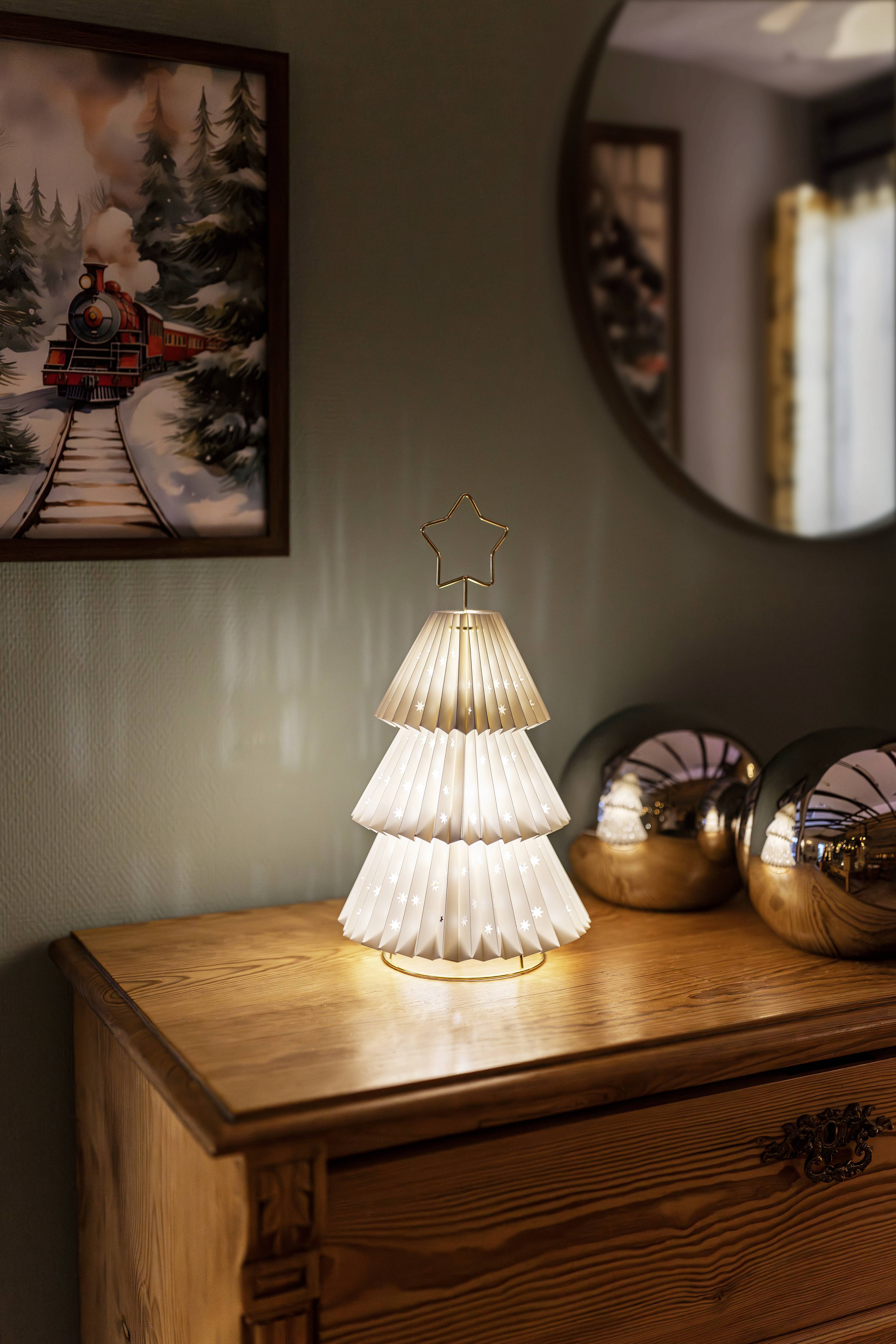 Une lampe décorative en forme de sapin de Noël avec une étoile au sommet est posée sur une commode en bois. À côté se trouvent des sphères argentées et une peinture représentant un train.