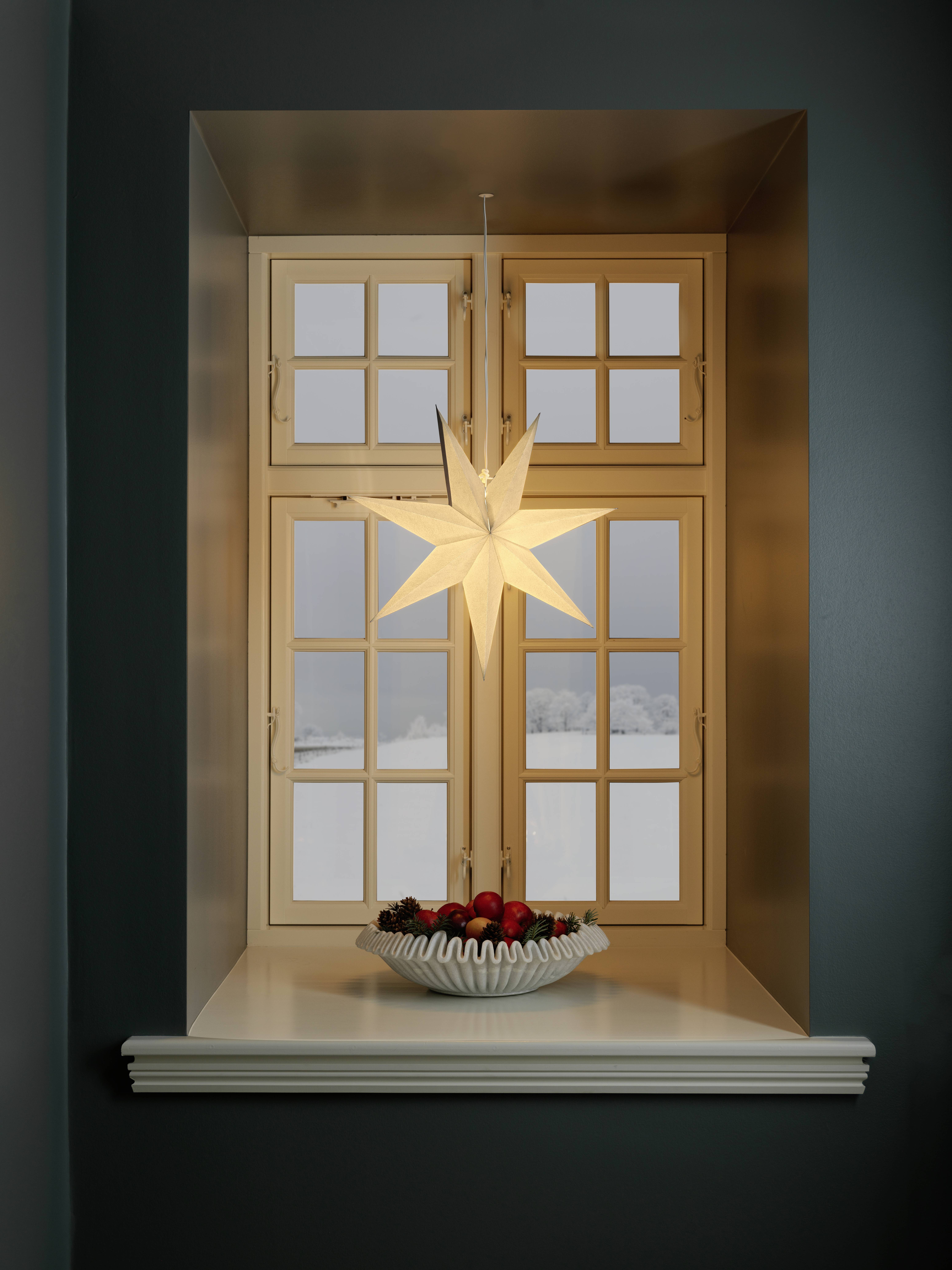 Une décoration en forme d'étoile illuminée est suspendue devant une fenêtre où la neige est visible à l'extérieur. Un bol rempli d'ornements colorés est posé sur le rebord de la fenêtre.