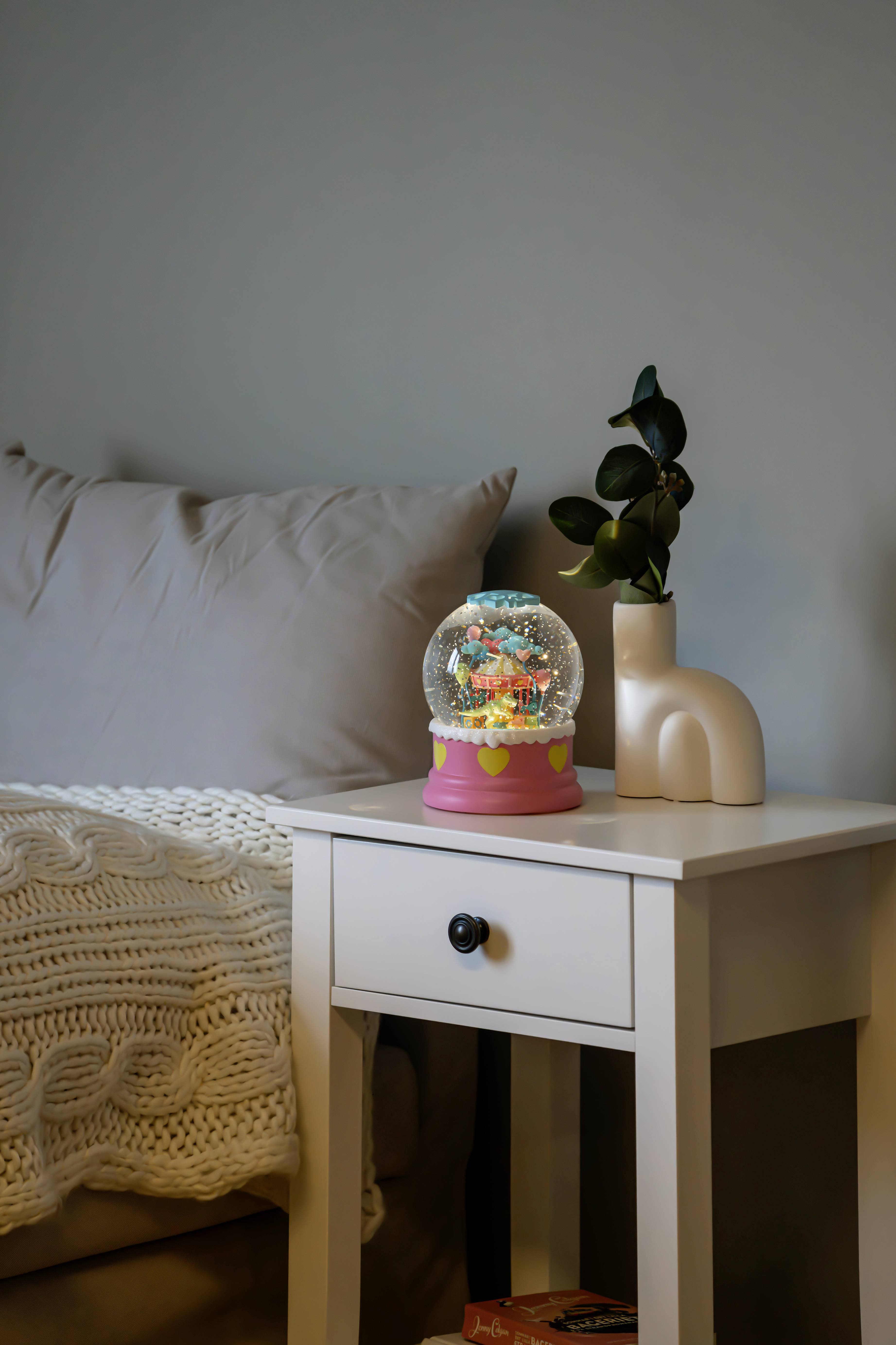 Un globe de neige orné de décorations colorées est posé sur une table d'appoint blanche, à côté d'un canapé gris sur lequel est posée une couverture tricotée, avec une plante verte dans un vase.