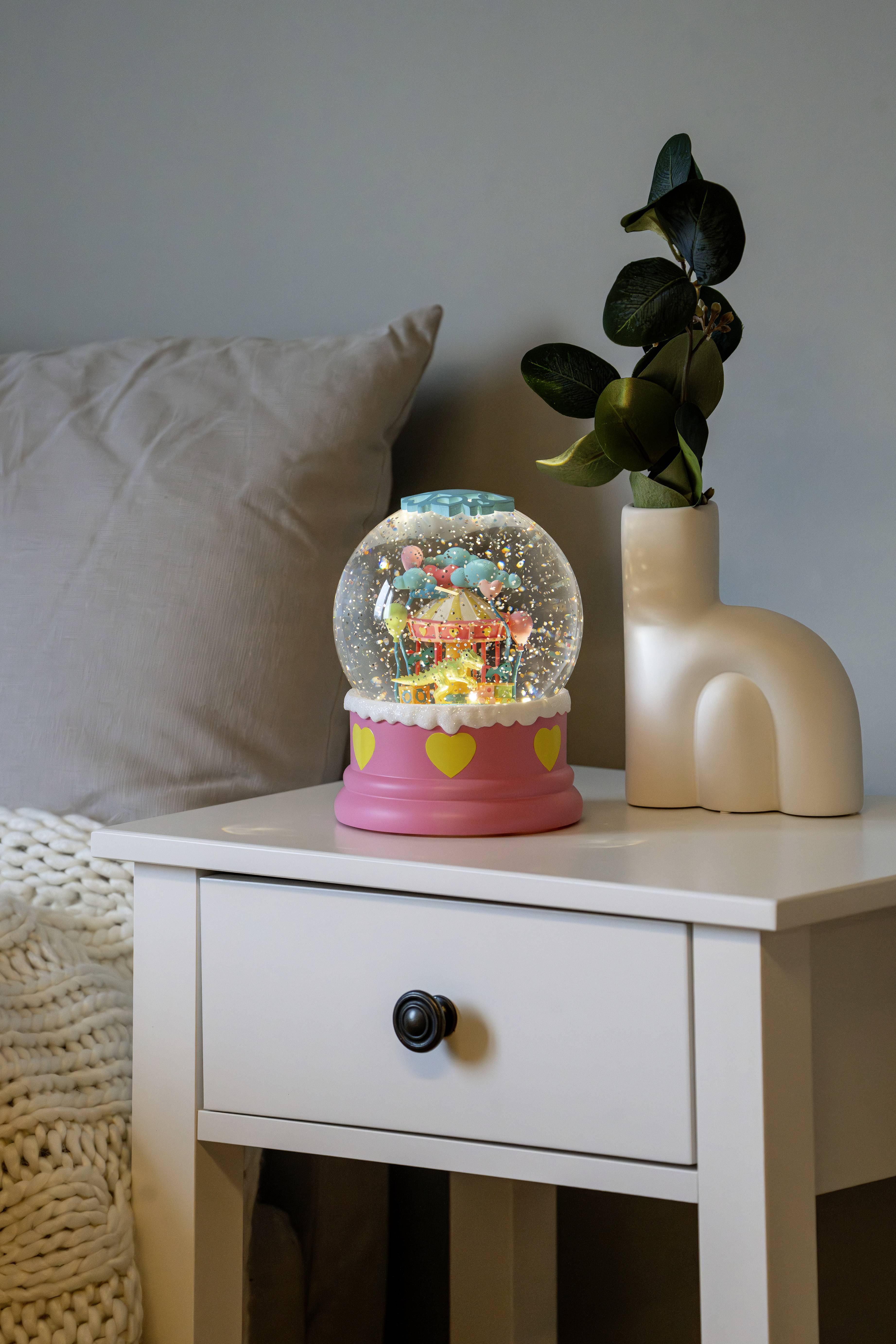 Un globe de neige décoré de couleurs vives est posé sur une table de chevet blanche, à côté d'un vase contenant des feuilles vertes, créant une ambiance chaleureuse.