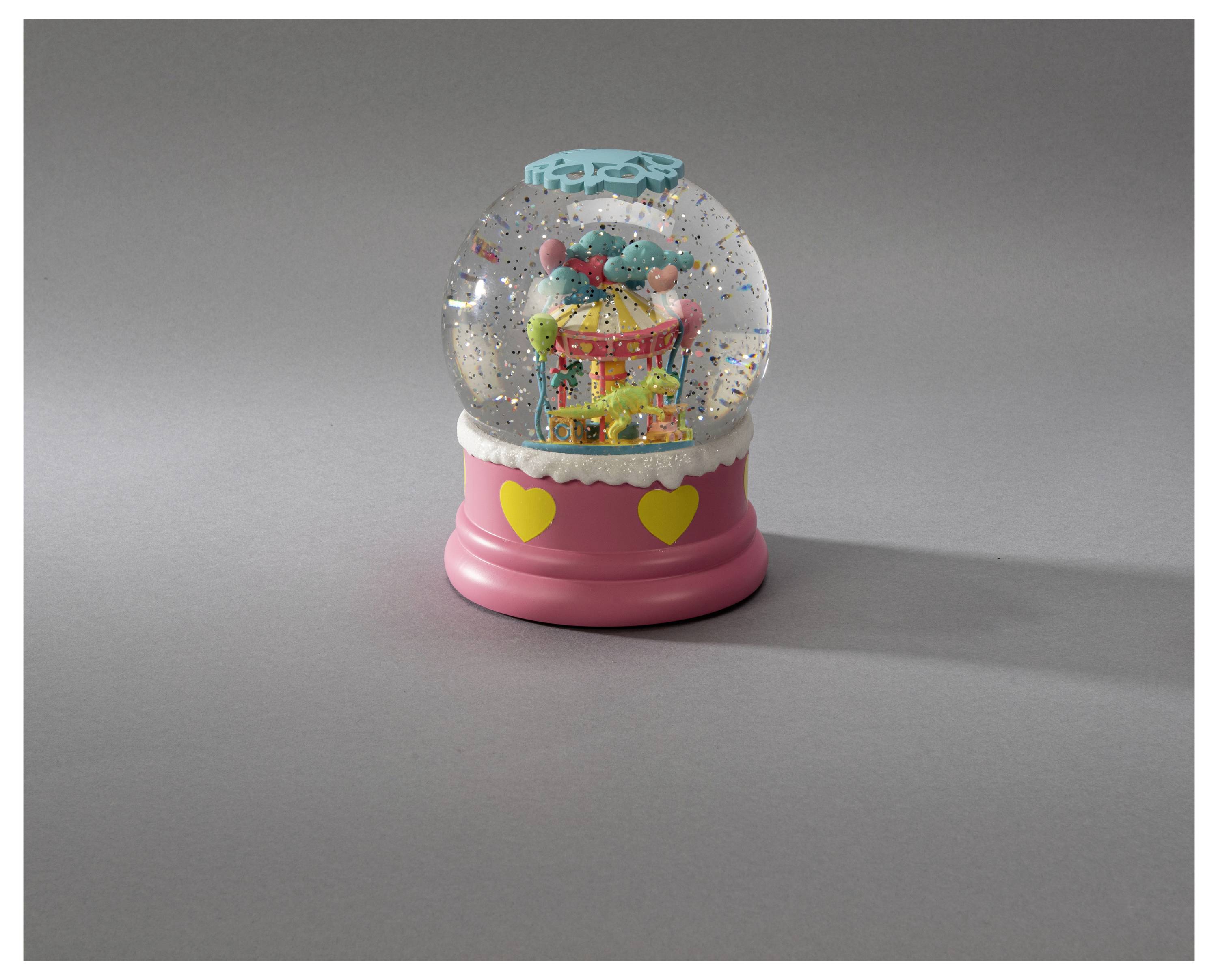 Un globe de neige avec une base rose ornée de cœurs jaunes, contenant un carrousel coloré avec des chevaux à l'intérieur, repose sur une surface grise.