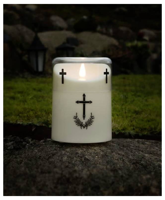 Une bougie blanche allumée avec des motifs de croix noirs, posée sur un rocher, entourée d'herbe et de pierres.