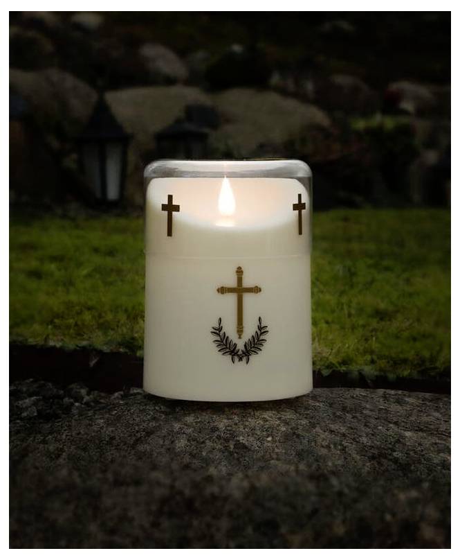 Une bougie blanche allumée avec des motifs de croix dorés est posée sur un rocher dans une zone herbeuse, symbolisant le souvenir ou la prière.