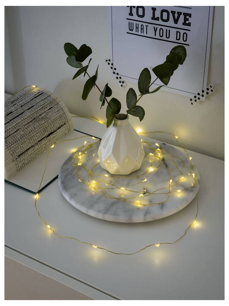 Un vase blanc orné de feuilles vertes est posé sur un plateau de marbre, entouré de guirlandes lumineuses, créant une atmosphère chaleureuse et accueillante.