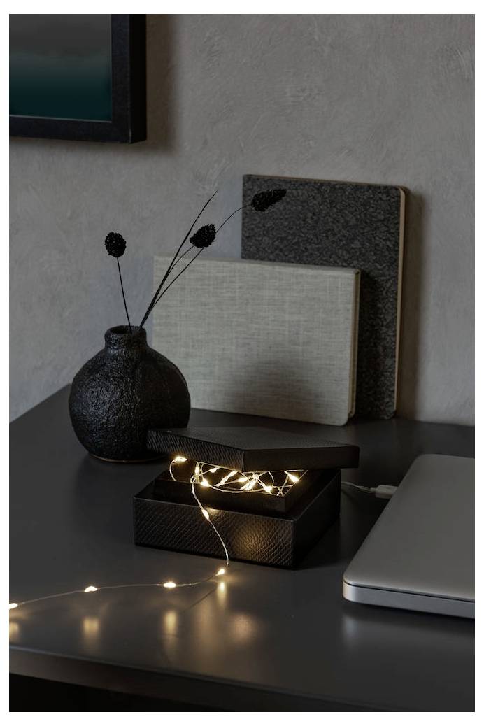 Un bureau avec un ordinateur portable, une boîte ouverte contenant des guirlandes lumineuses, deux livres fermés et un vase texturé tenant des plantes séchées.