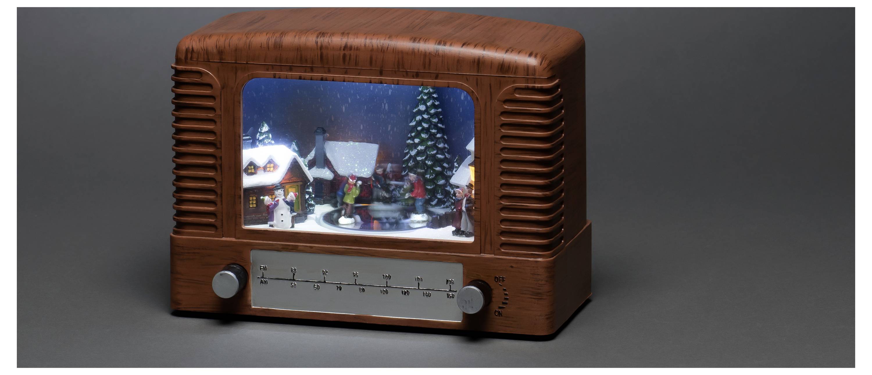 Un poste de radio de style vintage avec une scène de village d'hiver à l'intérieur de l'écran, présentant de la neige, des maisons et des figurines, sur un fond sombre.